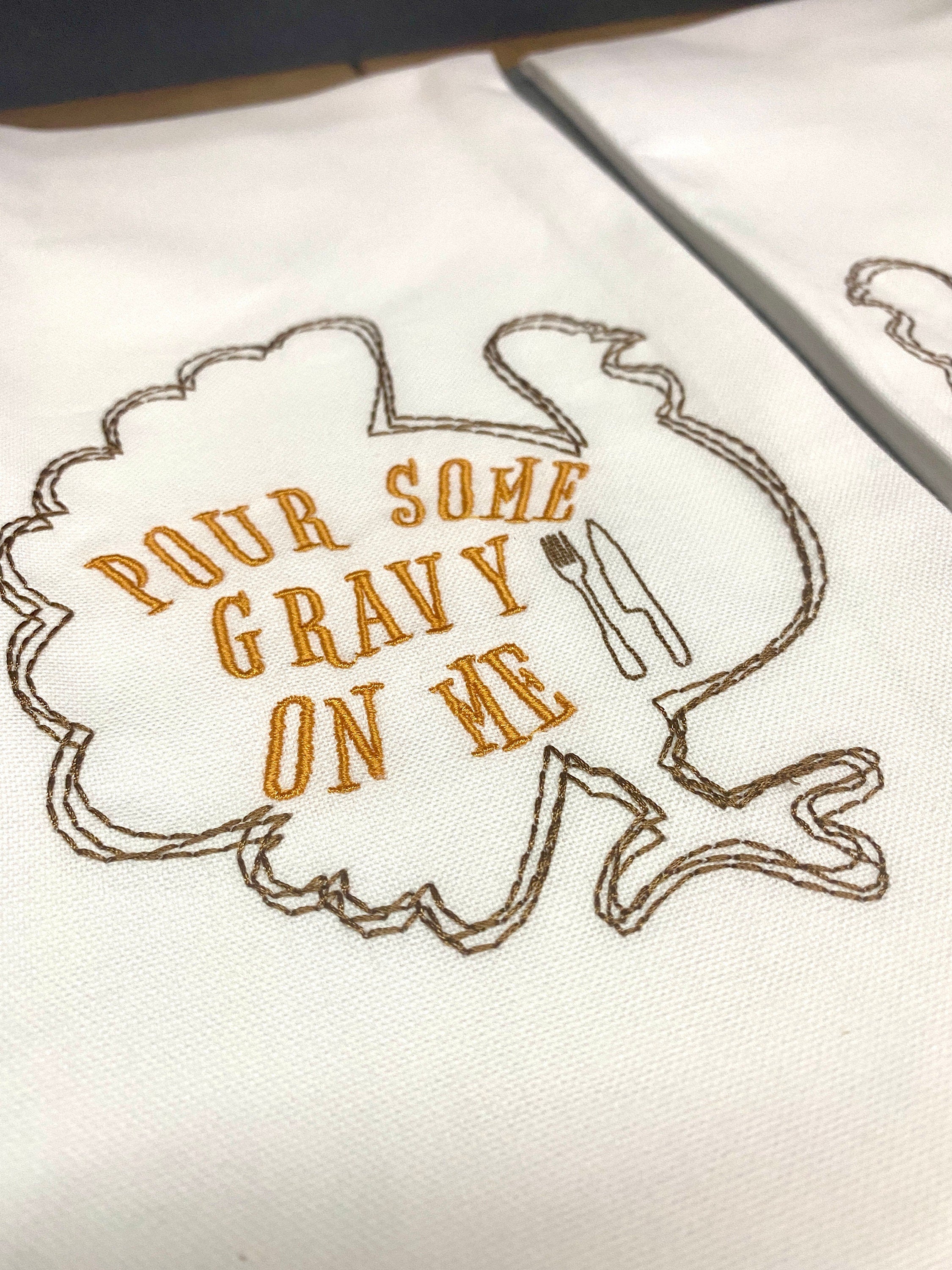 A white tea towel with the outline of a brown turkey embroidered with the words inside of the turkey: Pour Some Gravy on Me.