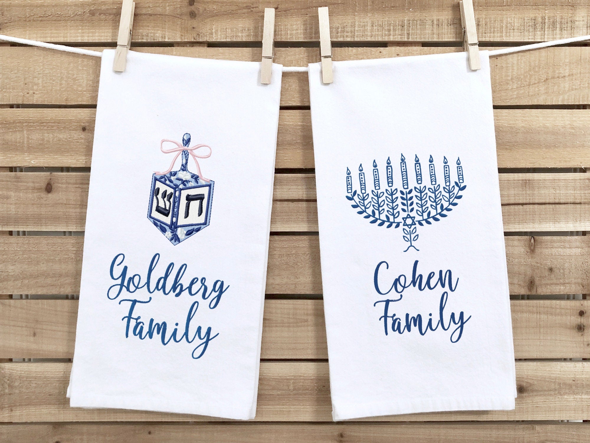 A photo of two white tea towel one with a dreidel in different shades of blue with a pink bow embroidered on a white cotton tea towel and one with a menorah in different shades of blue on a white cotton tea towel. Both are personalized with custom family names. They are set on a wood slatted background.