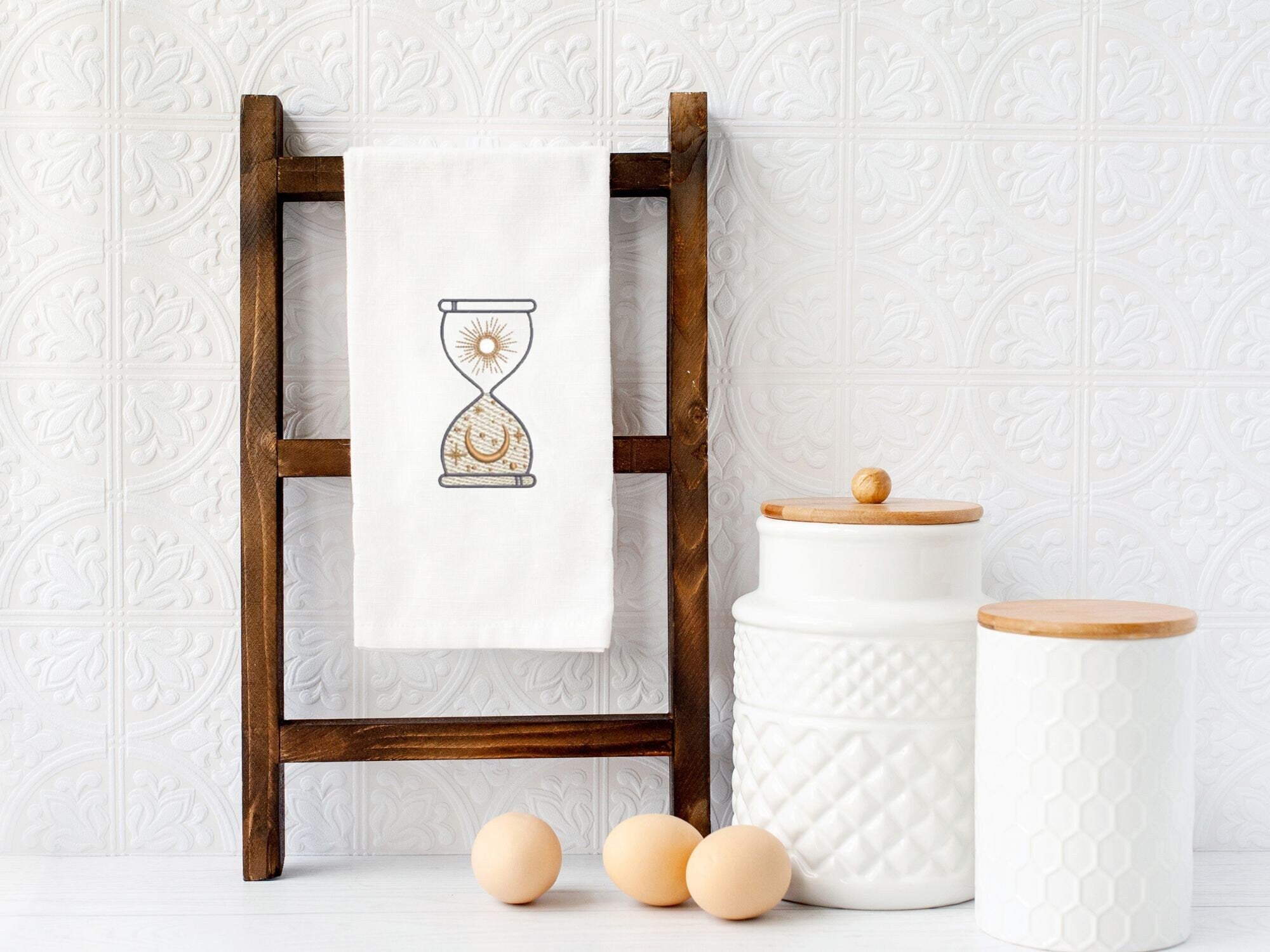 A white kitchen towel with a hourglass design embroidered on it. The hourglass is filled with a sun and moon design. The towel is hanging on a wooden ladder shelf with two white ceramic canisters with wooden lids on the bottom shelf.