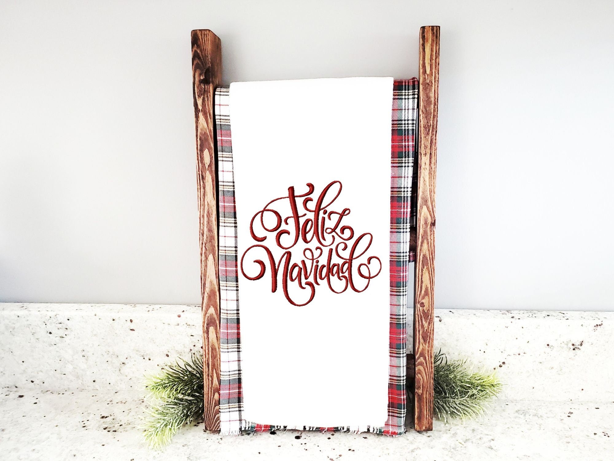 A white kitchen towel with red embroidered text that reads 'Feliz Navidad' displayed on a wooden stand with a red and black plaid fabric background.