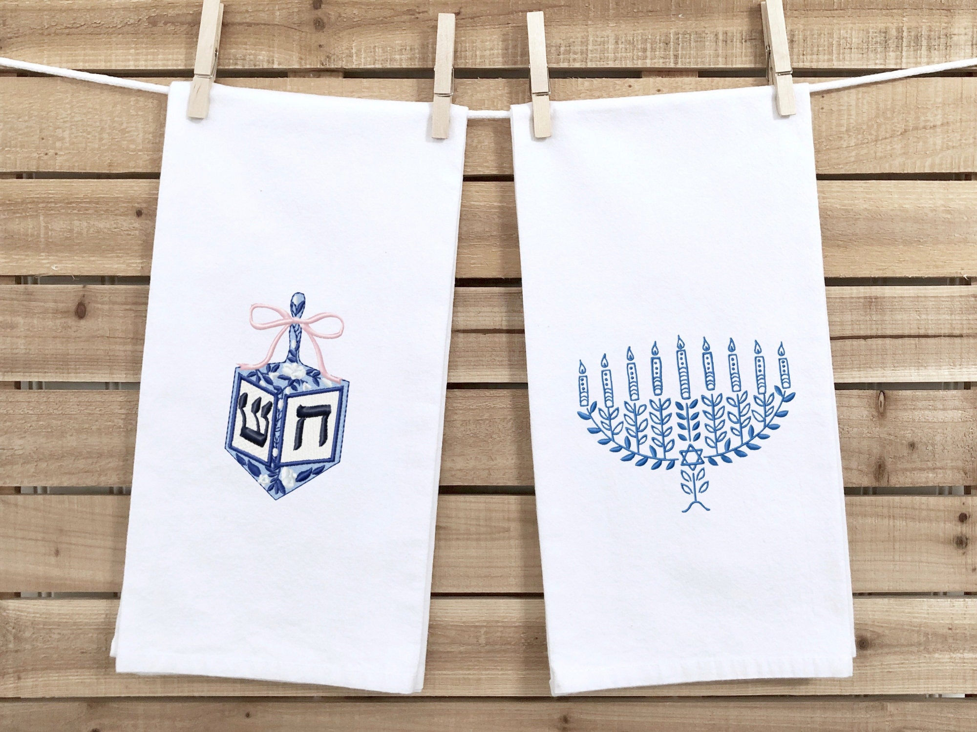 A photo of two white tea towel one with a dreidel in different shades of blue with a pink bow embroidered on a white cotton tea towel and one with a menorah in different shades of blue on a white cotton tea towel. This is showing both towels together with no personalization. The listing is for one towel - with the dreidel with no personalization, but the customer can choose both in the variations section.