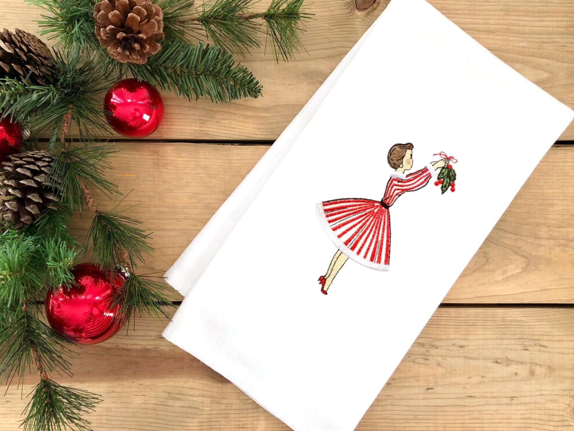 Image of 1950s inspired holiday decor tea towel of a mid century modern woman in a red and white striped dress hanging mistletoe. The tea towel is set on a wooden table with holiday greens and red Christmas ornaments.