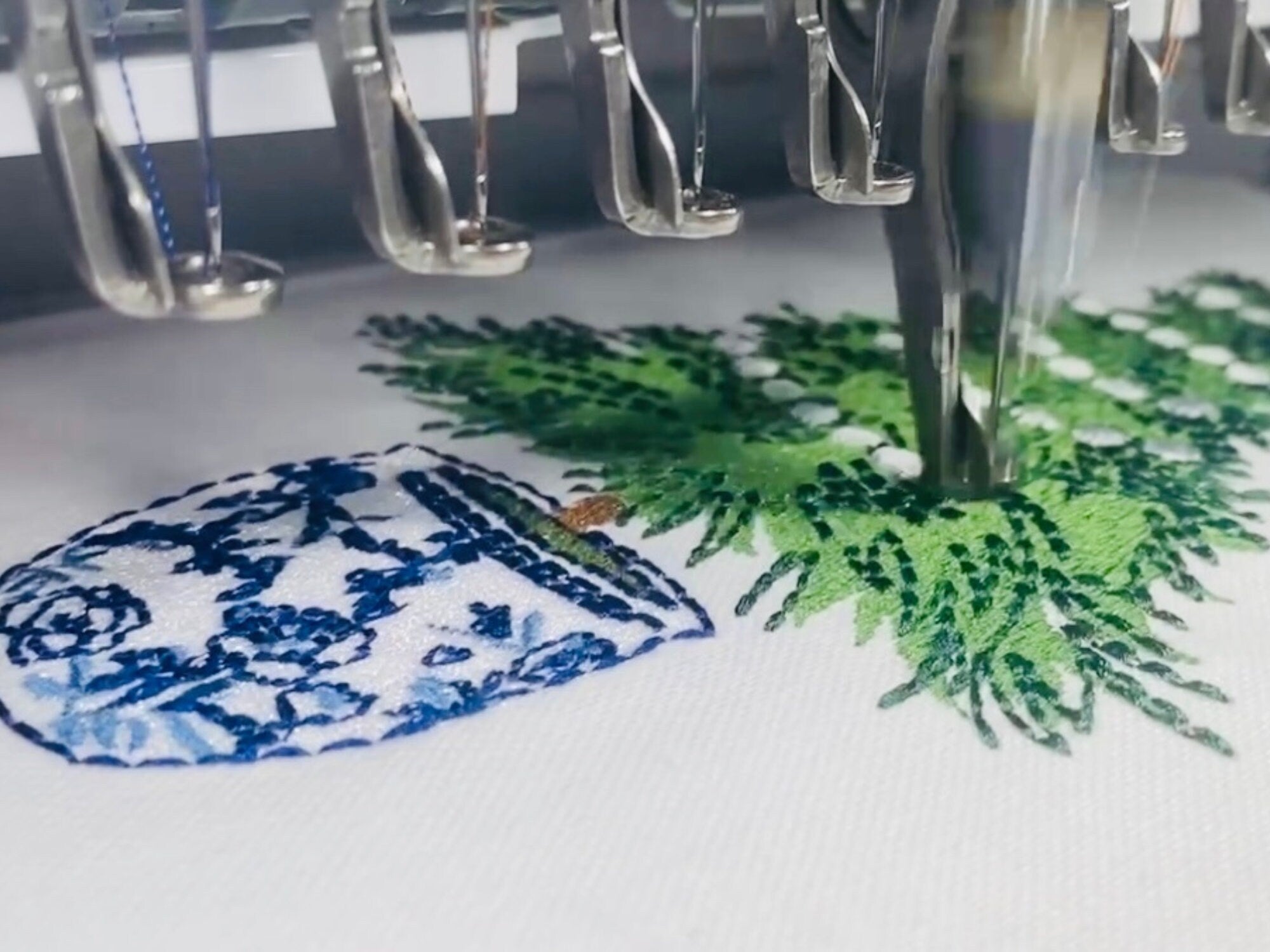 A close-up of an embroidery machine stitching a blue and white floral design into a white fabric. The design is of a potted plant with green leaves. The machine's needle is visible in the center of the image.