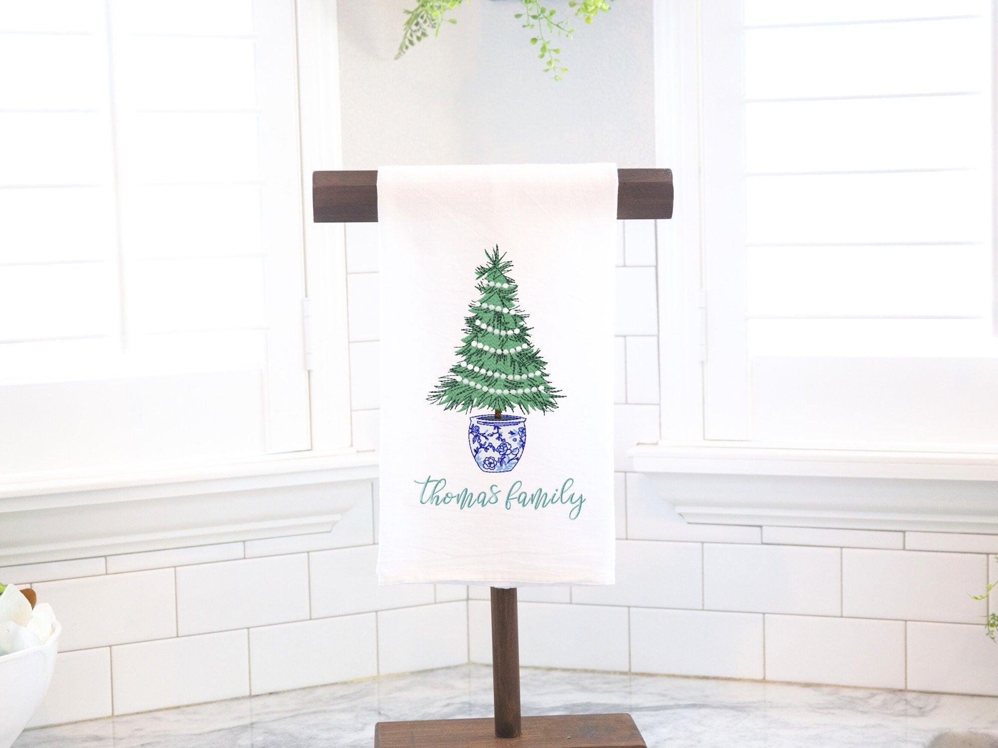 A white kitchen towel with a green Christmas tree in a blue and white pot. The tree is decorated with white lights. The towel has the text 'Thomas family' in green script.