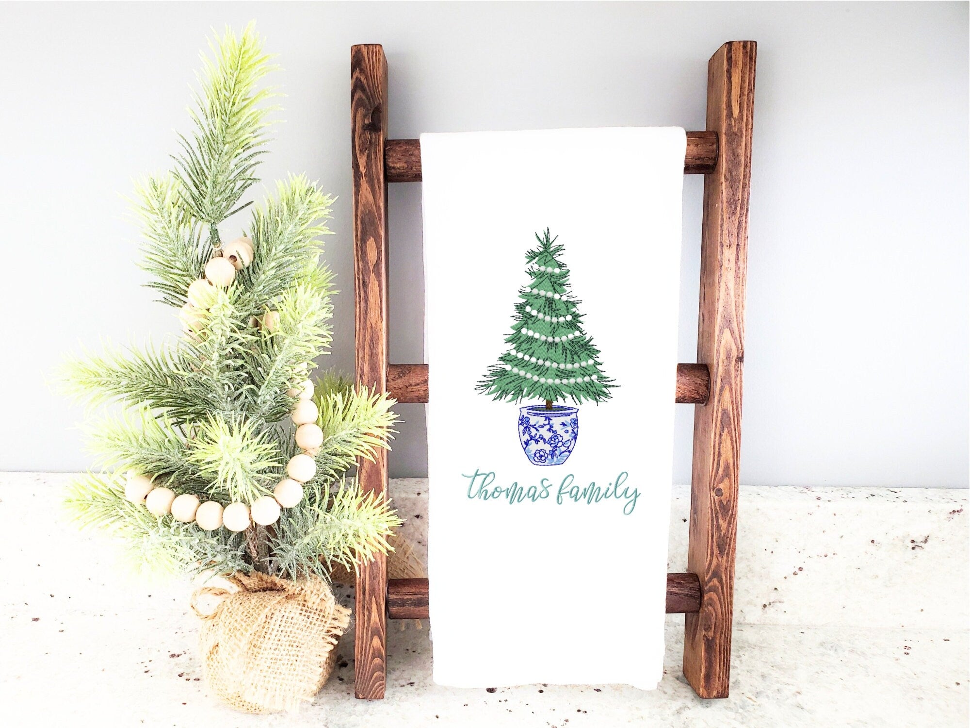 A white kitchen towel with a green Christmas tree in a blue and white chinoiserie planter. The tree is decorated with white lights. The text 'Thomas family' is embroidered below the tree.