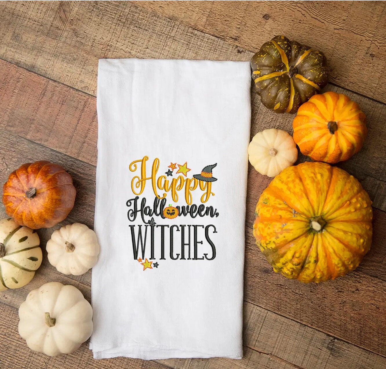White tea towel with Happy Halloween Witches embroidered set in the middle of different sized pumpkins and gourds.