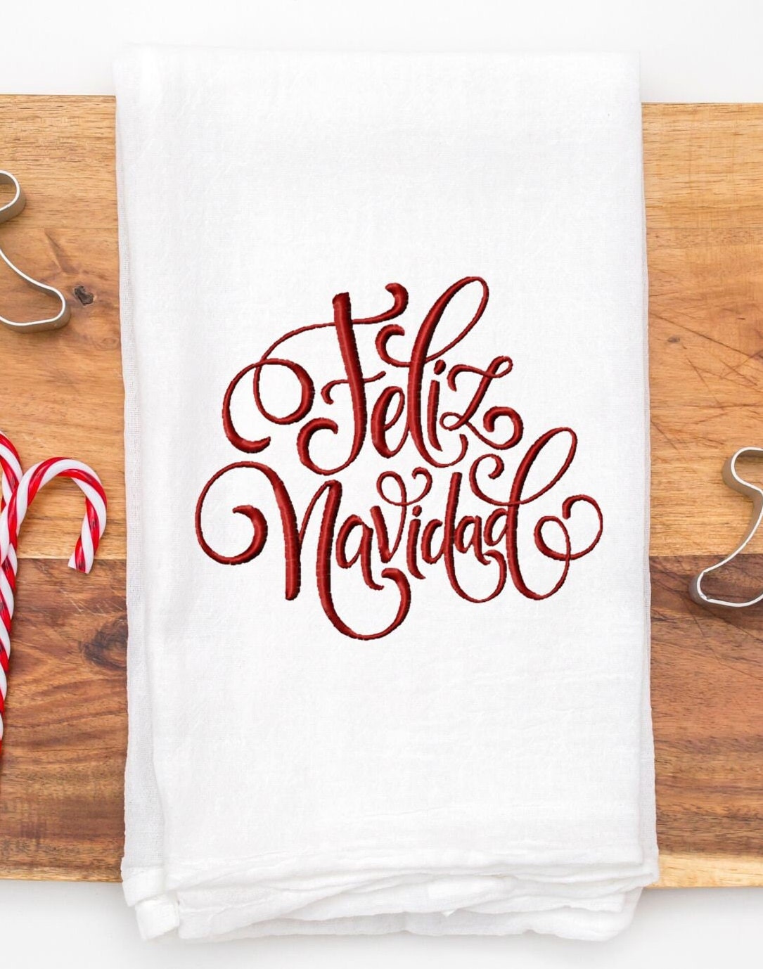 A white kitchen towel with red embroidered text that reads 'Feliz Navidad' set on a cutting board with a candy cane to the left.