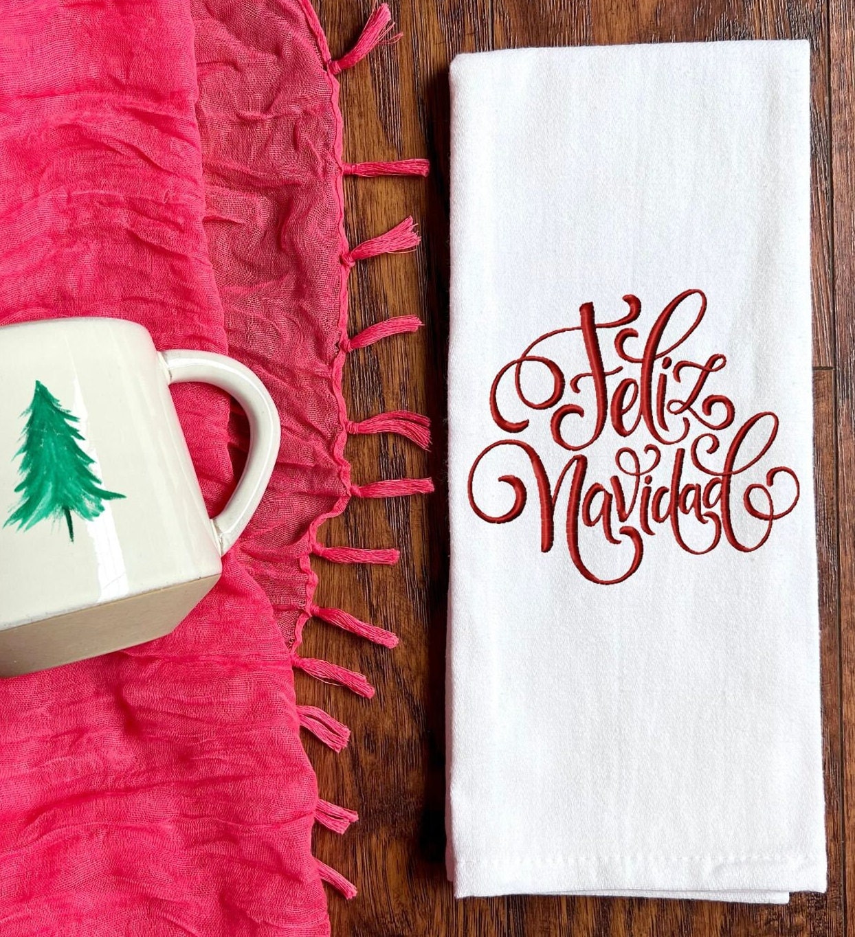 White kitchen towel with red embroidered text that says 'Feliz Navidad' set on a brown table with a red fringed table throw and a mug with a Christmas tree next to it.