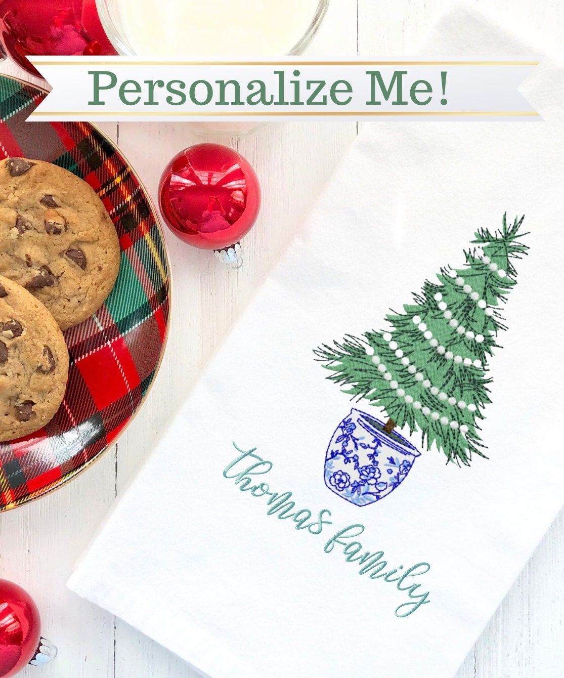 A white kitchen towel with a green Christmas tree in a blue and white chinoiserie pot. The tree is decorated with white ornaments. The text 'Thomas Family' is embroidered in green cursive below the tree. The towel is on a white wooden surface with red Christmas ornaments and cookies.