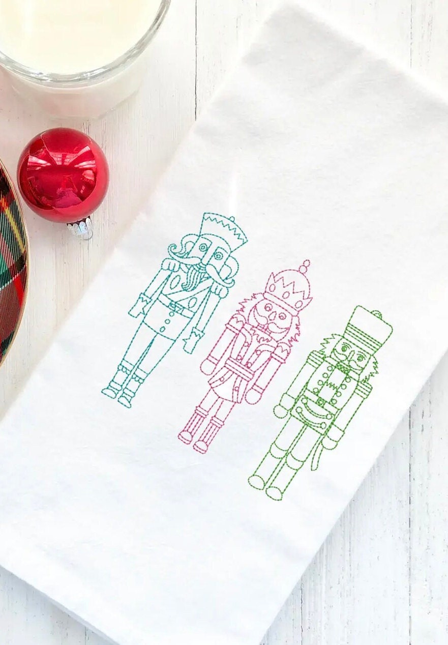 Three embroidered nutcracker figures on a white kitchen towel. The nutcrackers are outlined in green, pink, and blue thread. The nutcrackers are standing in a row and are facing the viewer.
