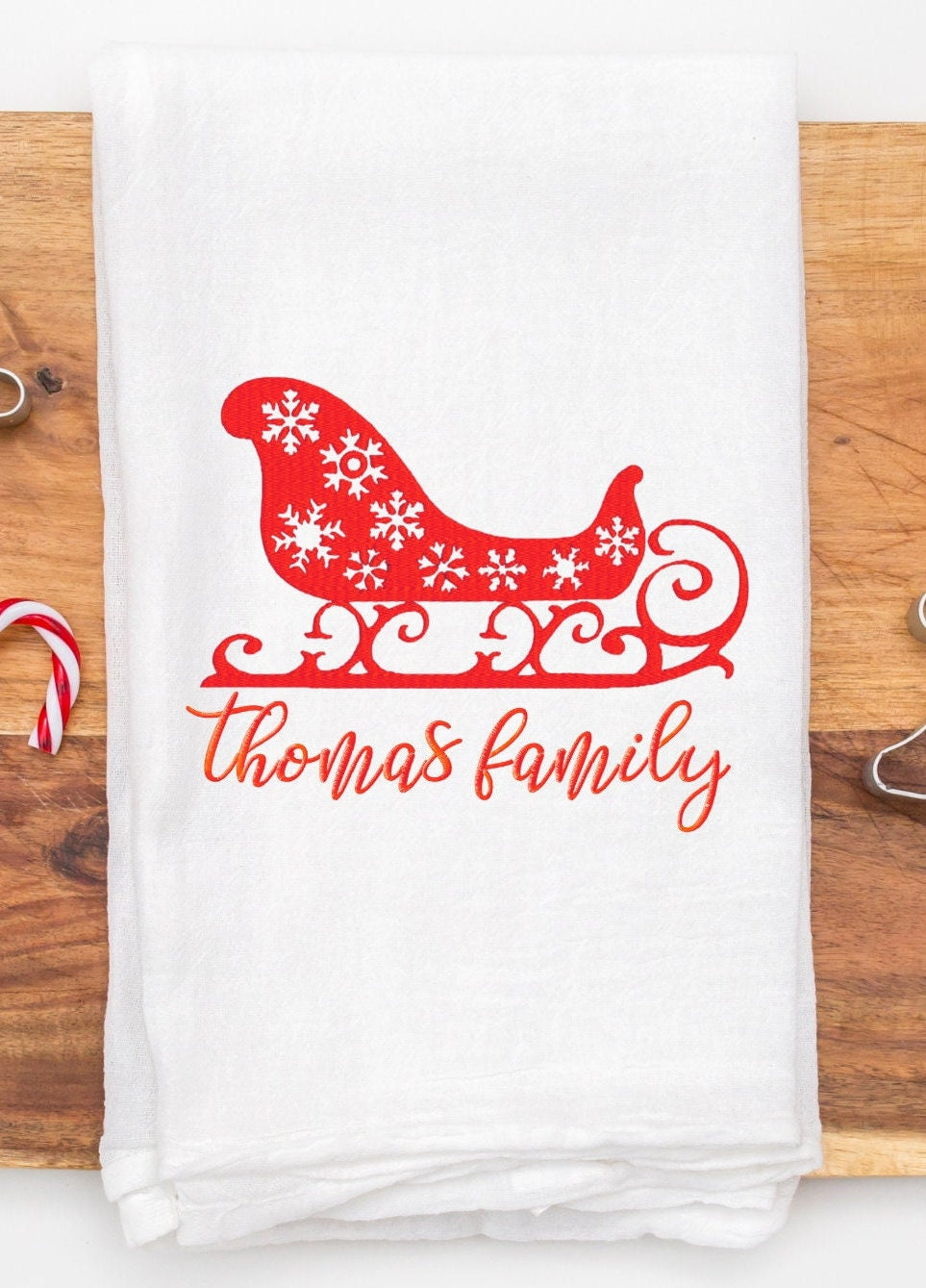 A white kitchen towel with a red Christmas sleigh design with white snowflakes. The text 'thomas family' is embroidered in red below the sleigh.