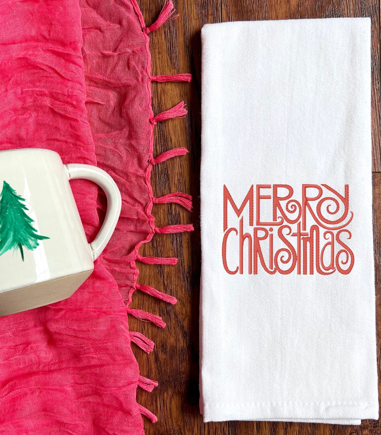 Merry Christmas embroidered in a pretty font where the R's and S's have a twirl at the end. Red embroidery. Set on a medium toned wooden table with a red scarf next to it and a white coffee mug with a simple, green painted pine tree on it.
