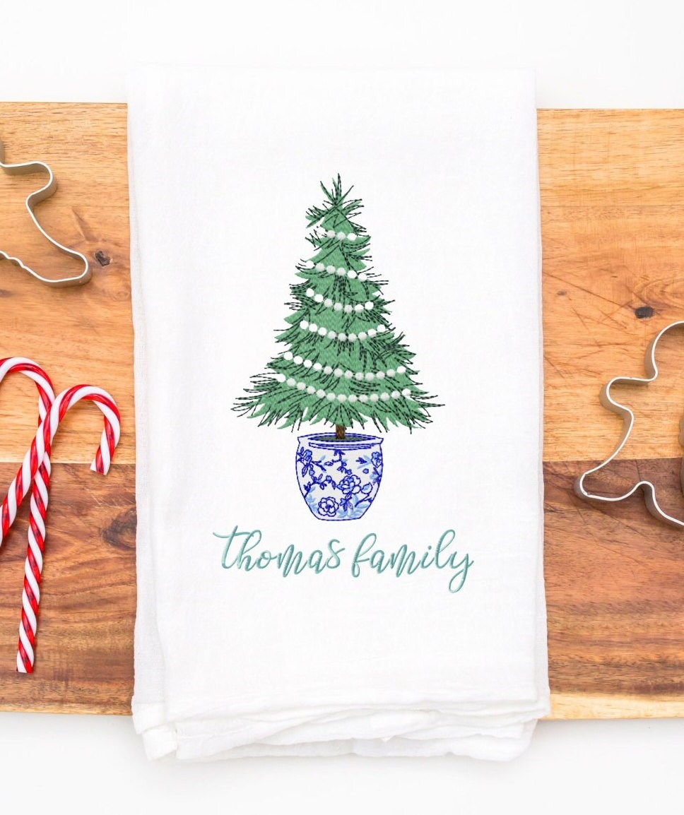 A white kitchen towel with a green Christmas tree in a blue and white patterned pot. The tree is decorated with white lights. The text 'thomas family' is embroidered below the tree.