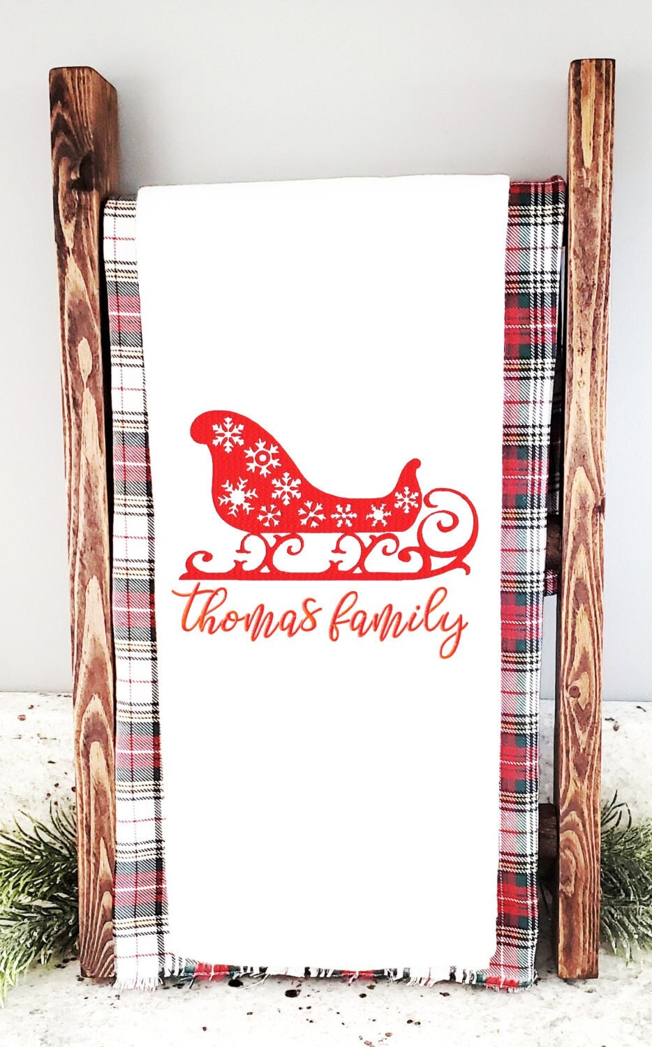 A white kitchen towel with a red sleigh with white snowflakes and embroidered with 'thomas family' in red.