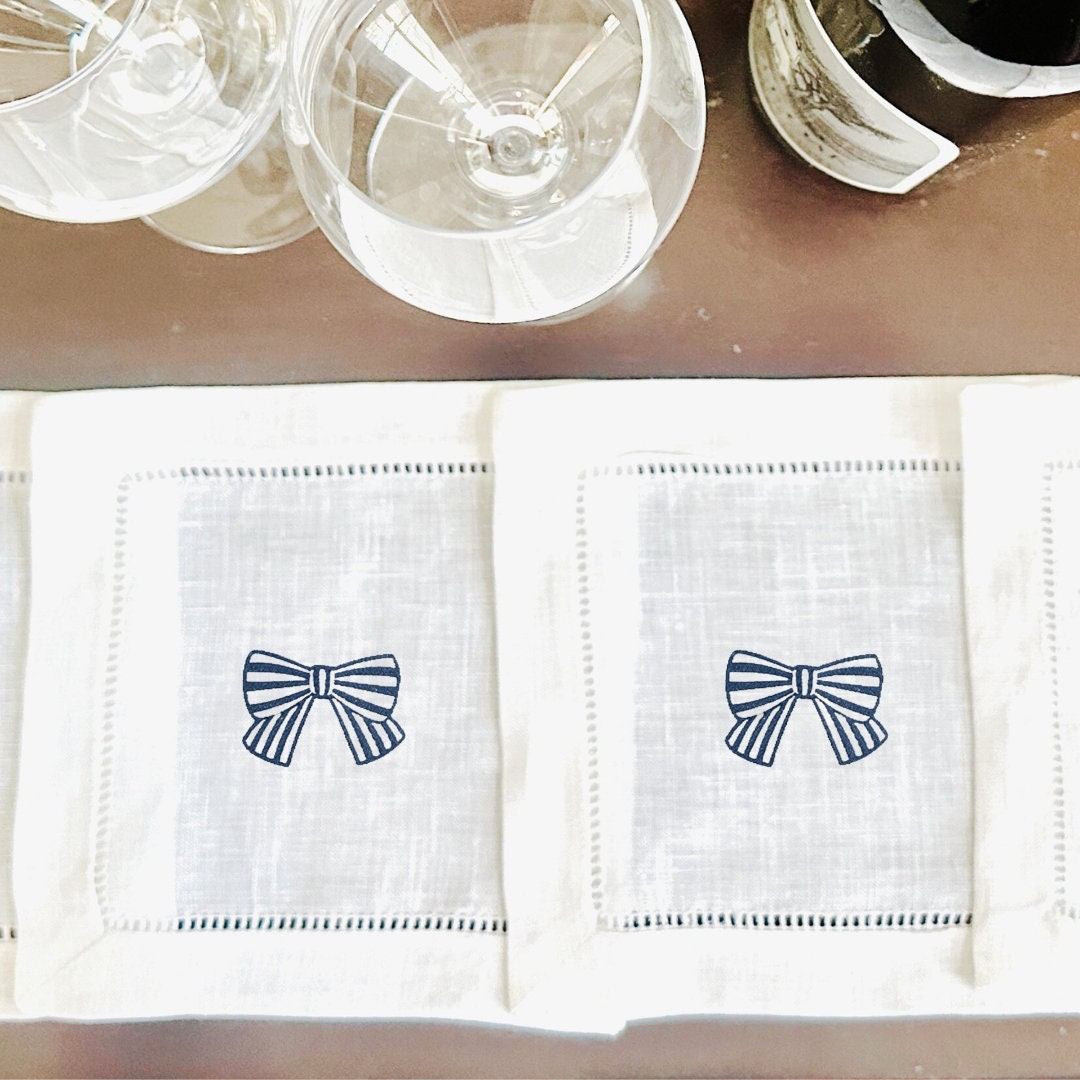 Two white linen napkins with embroidered blue and white bows set on a bar next to wine glasses.