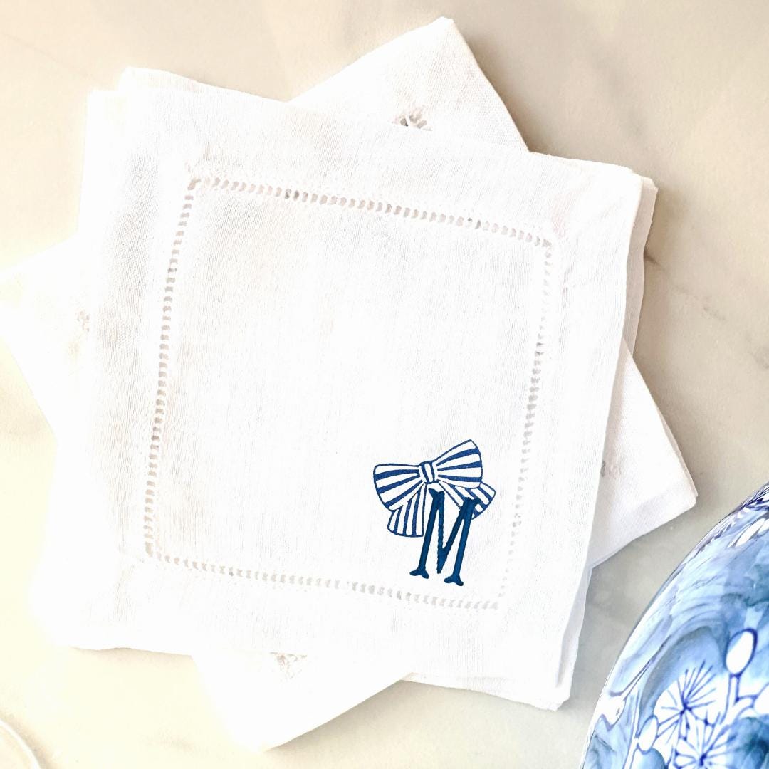 White linen napkins with a blue and white striped bow and the letter 'M' embroidered on them. Set on a marble counter with a chinoiserie blue and white vase on the lower right. Cocktail napkins are 6"x6" square.
