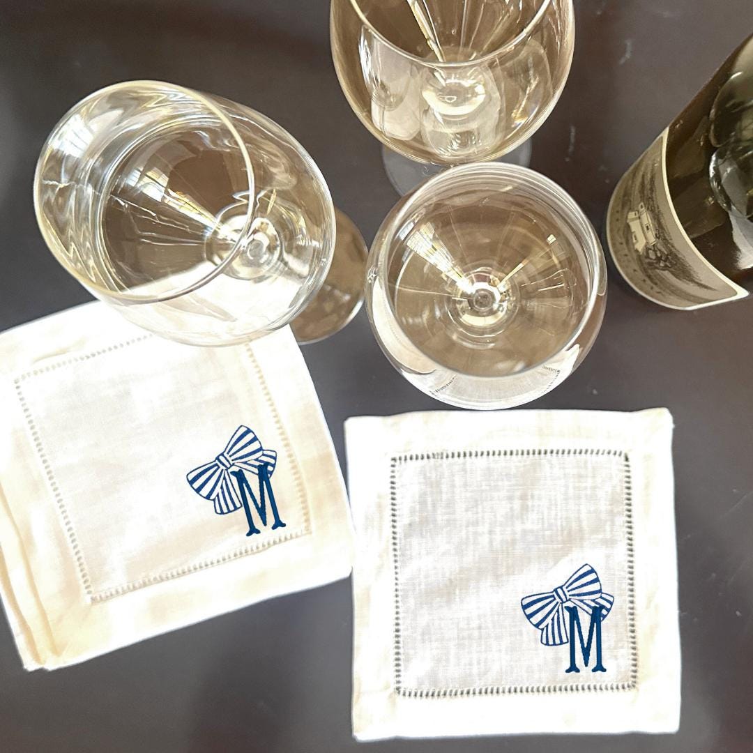 Two white linen cocktail napkins with blue and white striped bows and the letter 'M' embroidered on them. The napkins are on a dark brown surface with two clear wine glasses and a bottle of wine.