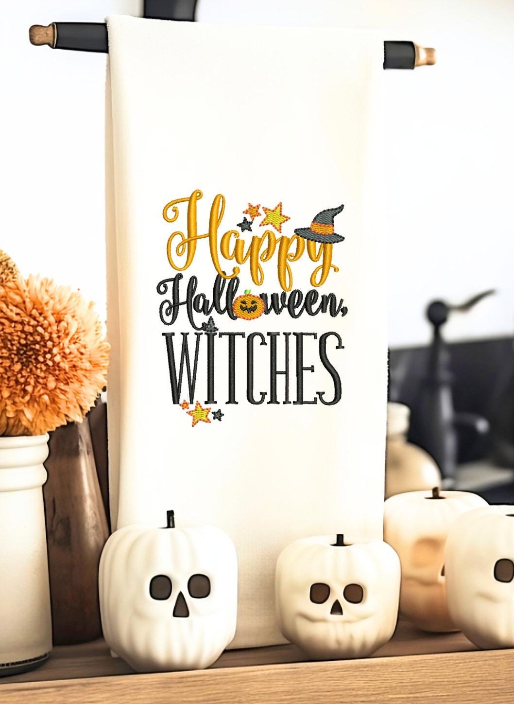 A bright white tea towel with the words Happy Halloween Witches embroidered on it hanging from a black tea towel stand with white pumpkin skulls next to orange mums in a white ceramic vase.