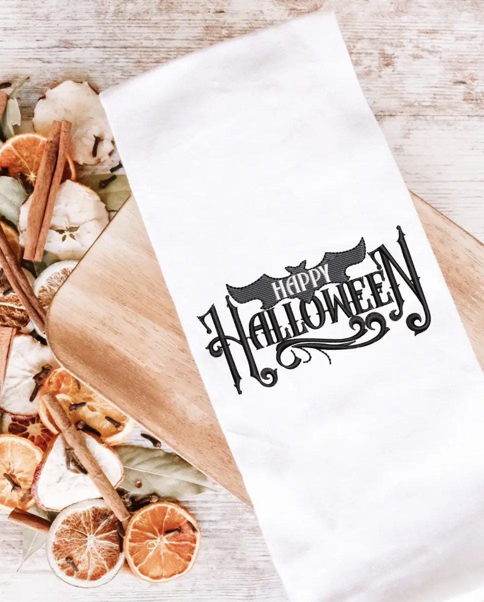 White tea towel with Happy Halloween on a cutting board with an apple, orange, cinnamon potpourri next to it. The stitching saying "Happy Halloween" is done all in black.