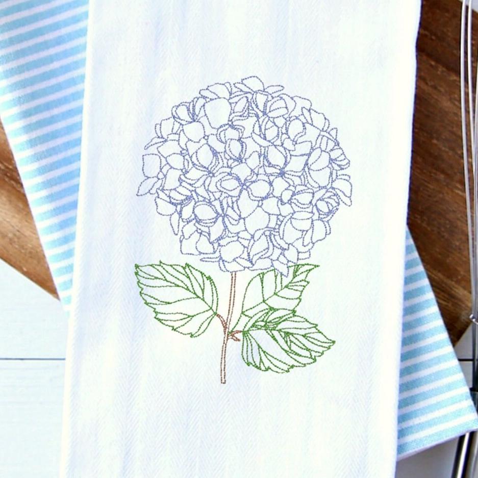 A beautiful hydrangea embroidered on a tea towel and set on a cutting board over a blue and white striped tea towel. Towels are approximately 17x30".
