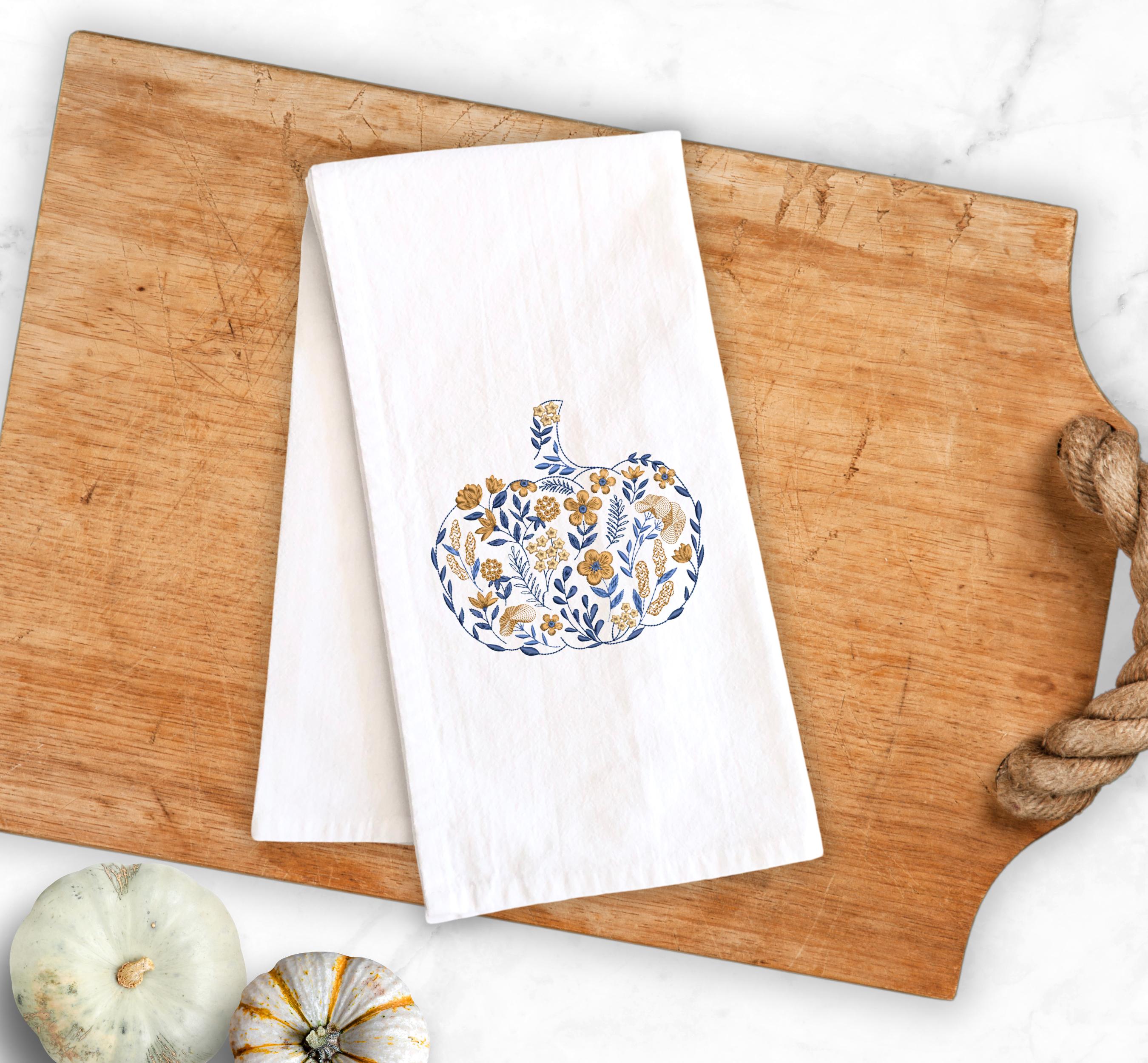 Autumnal home decor of a blue and golden orange Embroidered Pumpkin, on top of a cutting board with a rope handle and pumpkins to the lower left  used for Fall Hosting Decor, Thanksgiving Towel, Hostess Gifts, for Your Best Friend.
