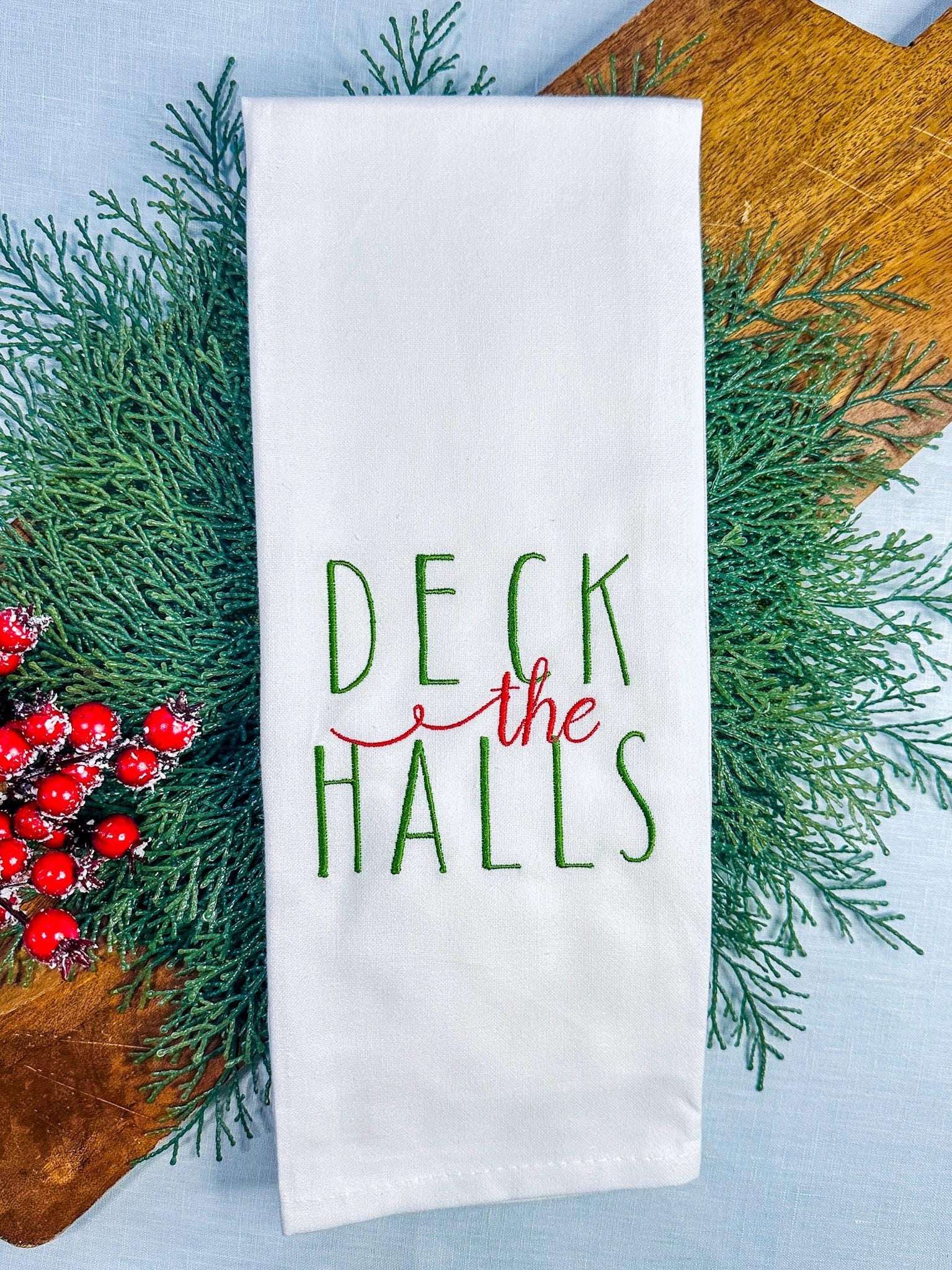 A white tea towel with Deck the Halls embroidered on it in green and red set on a cutting board with holiday greens and red Christmas berries.