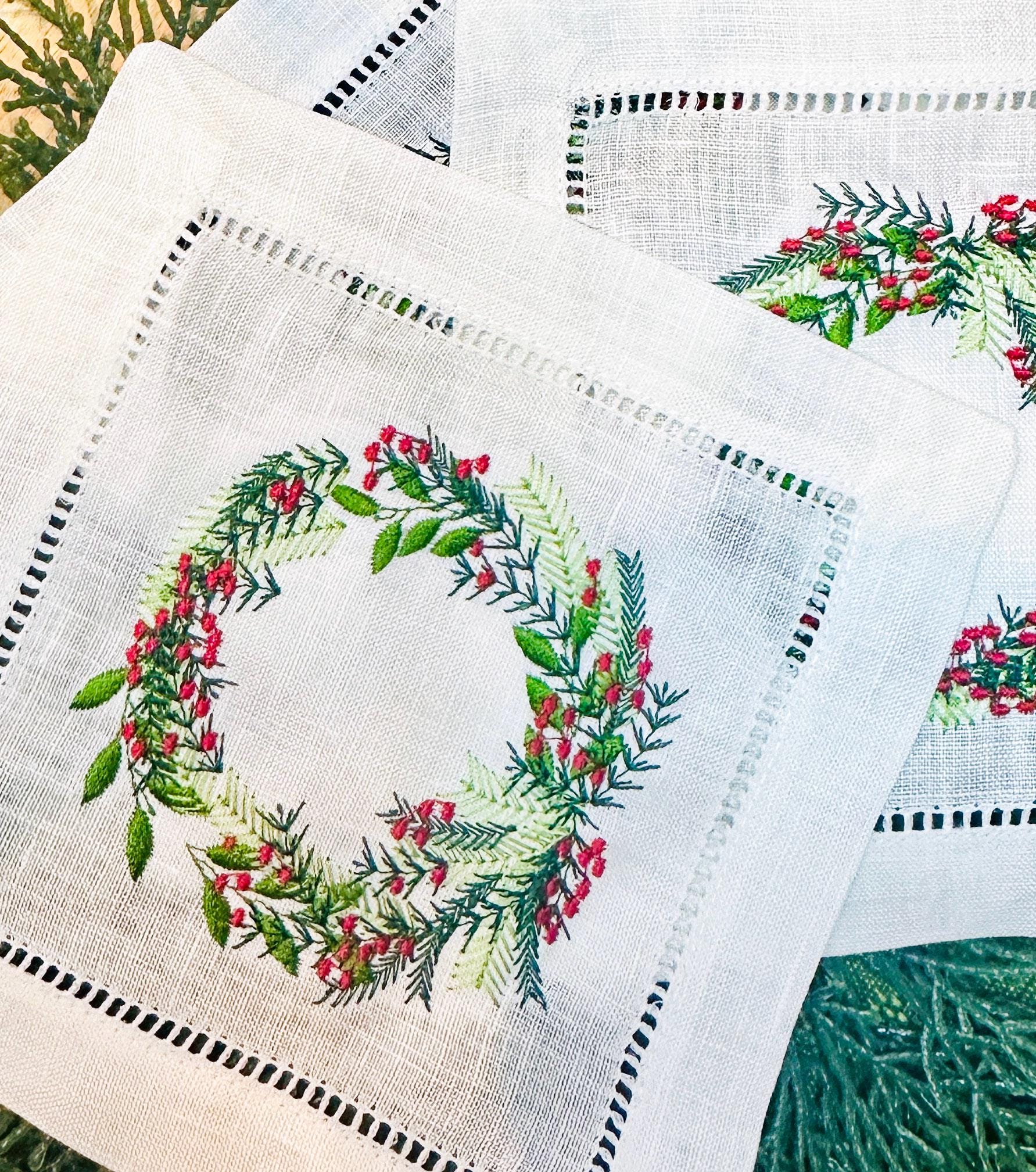 Christmas wreath with different color greens and red berries embroidered on a white cocktail napkin. Perfect gift for Mother in Law Christmas. There are three napkins on an evergreen placemat.