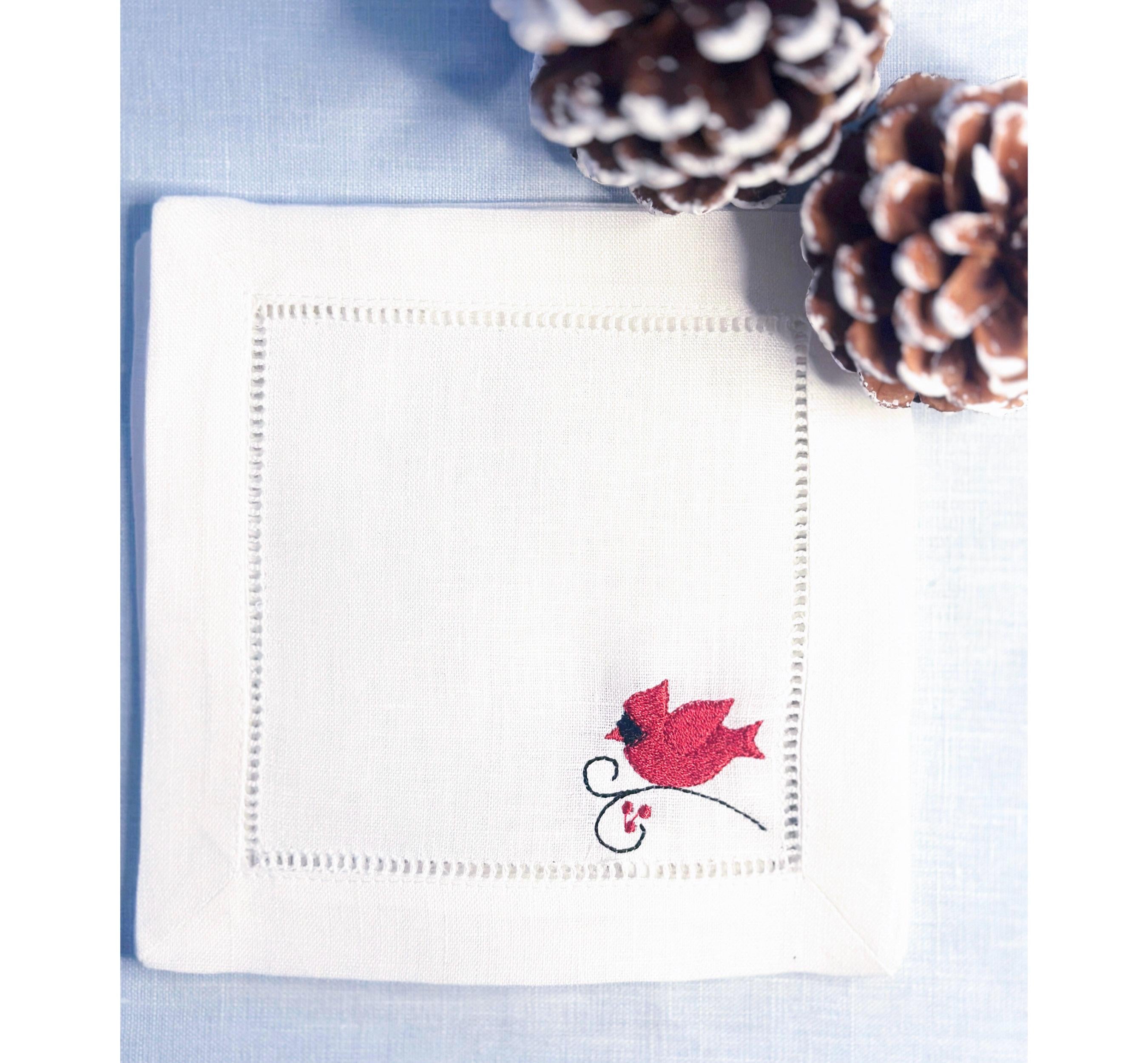 White linen cocktail napkin with a red male cardinal embroidered in the bottom right corner of the hemstitch napkin set on a light blue linen tablecloth with pine cones on top for bird watcher gift.