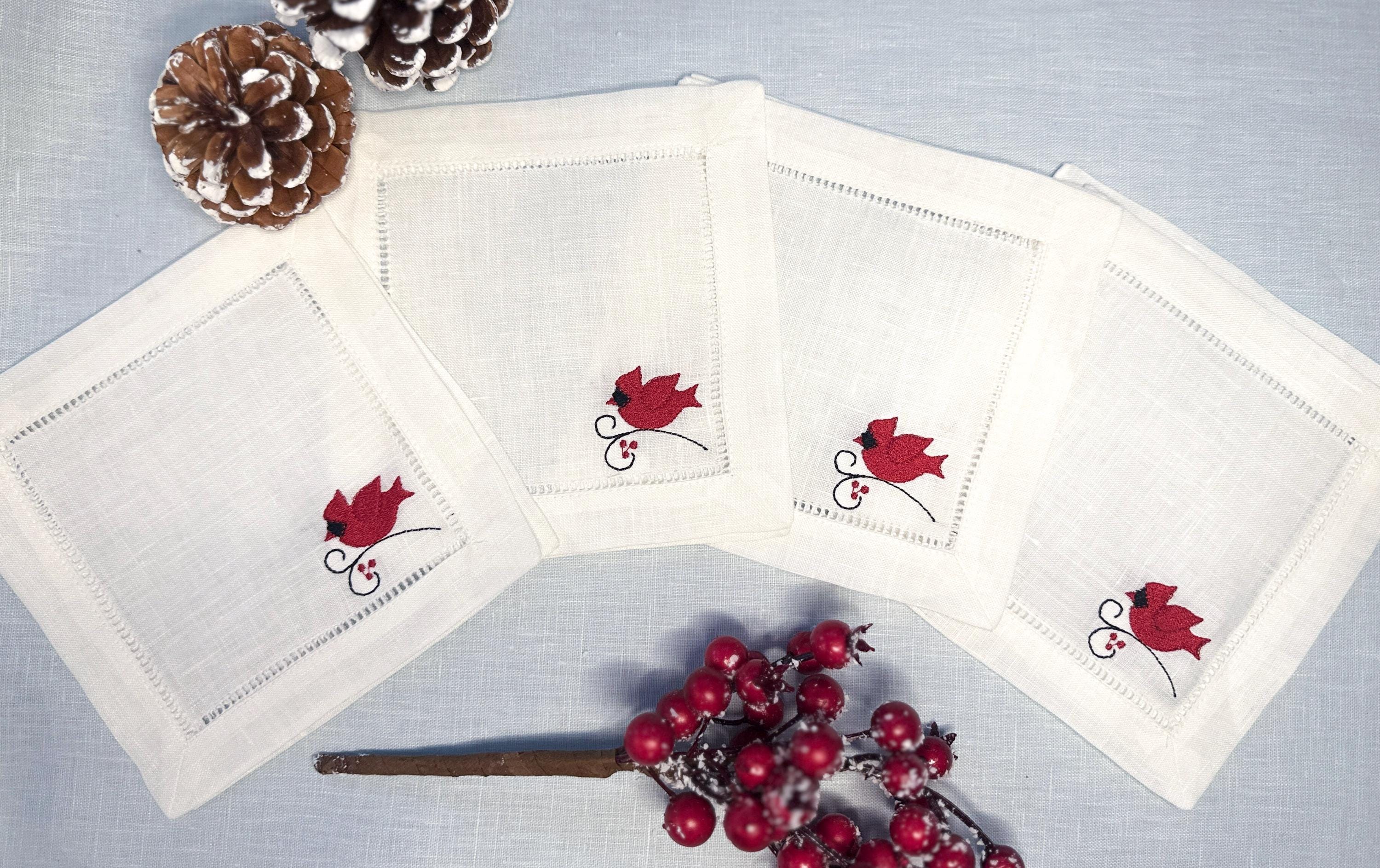 Set of 4 white linen cocktail napkins with a red male cardinal embroidered in the bottom right corner of the hemstitch napkin set on a light blue linen tablecloth with pine cones on top, and Christmas berries on the bottom - for bird lover gift.