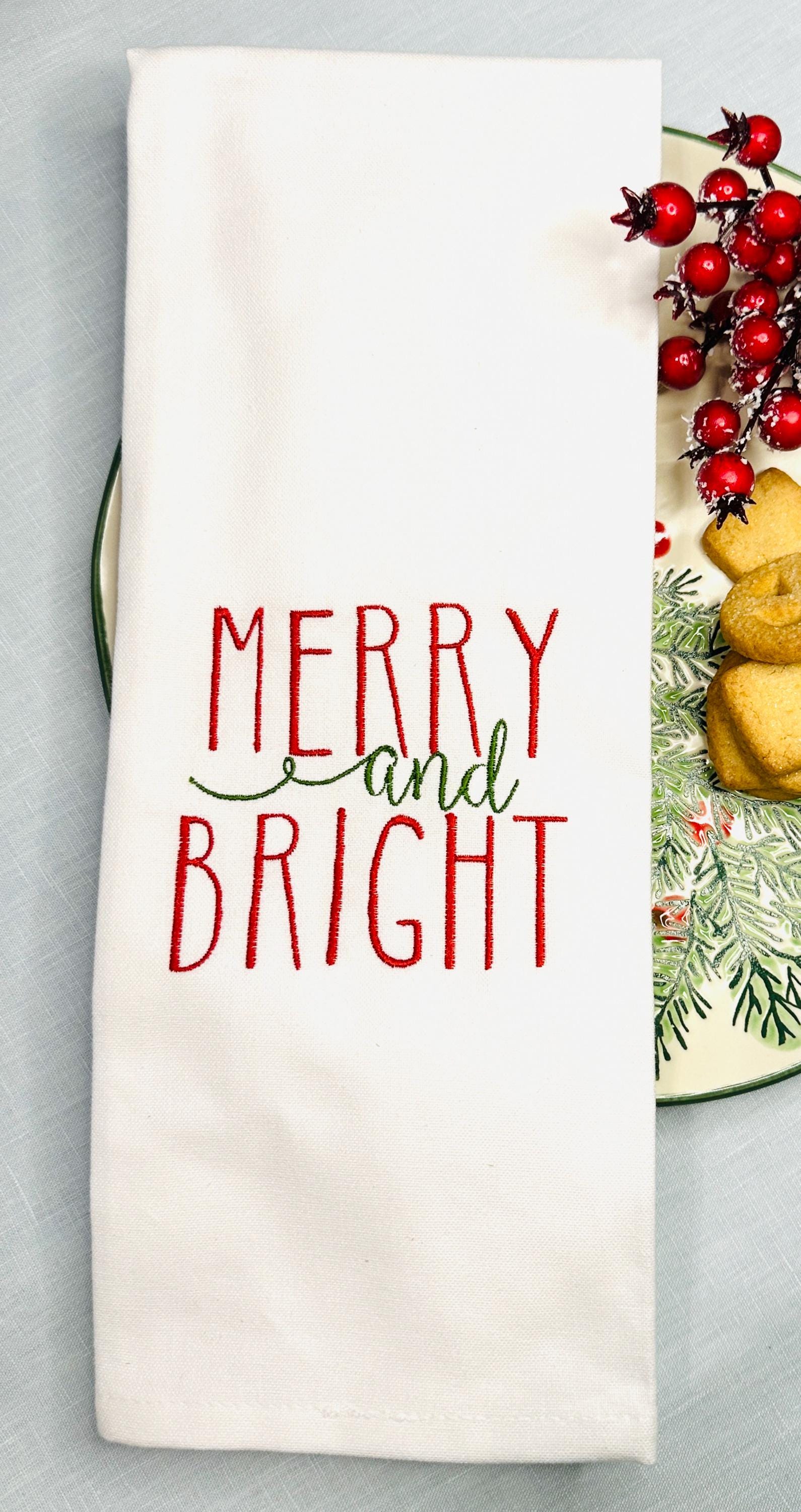 White tea towel embroidered with Merry and Bright in Green and red on top of a holiday plate with cookies and berry branches.