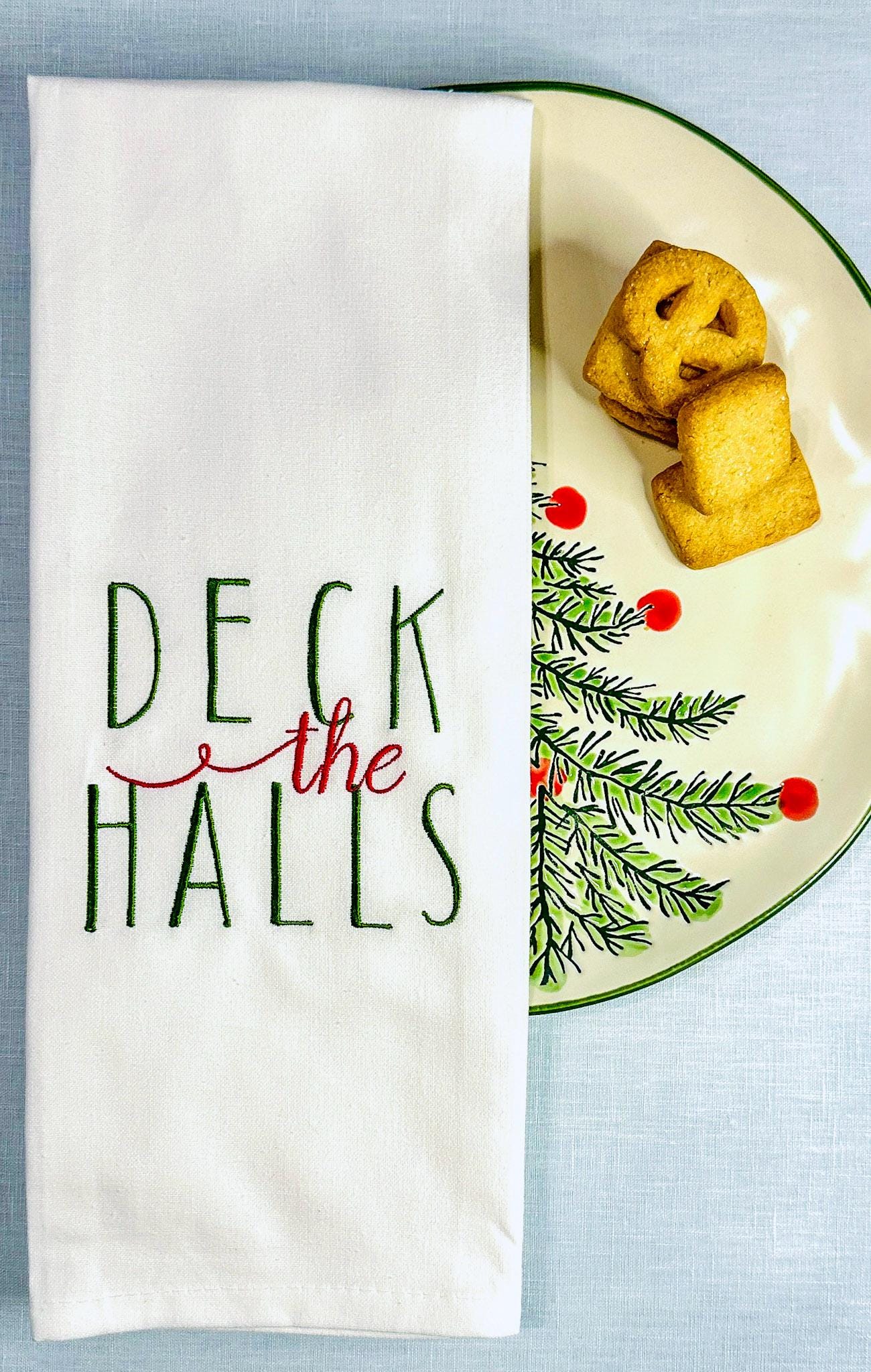 White tea towel with Deck the Halls embroidered in green and red set on a Christmas plate with a holiday tree and cookies.