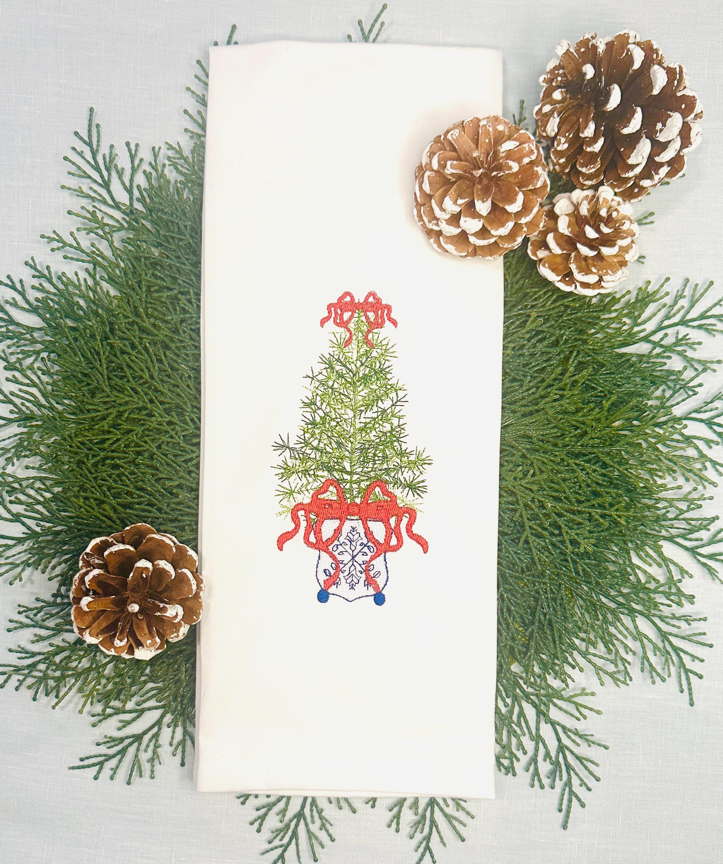 Simple, elegant chinoiserie style Christmas tree stitched on a white tea towel set upon an evergreen placemat with "snowy" acorns on top and bottom. There is a red bow on top and the bottom of the tree with blue chinoiserie stitching on a white pot.