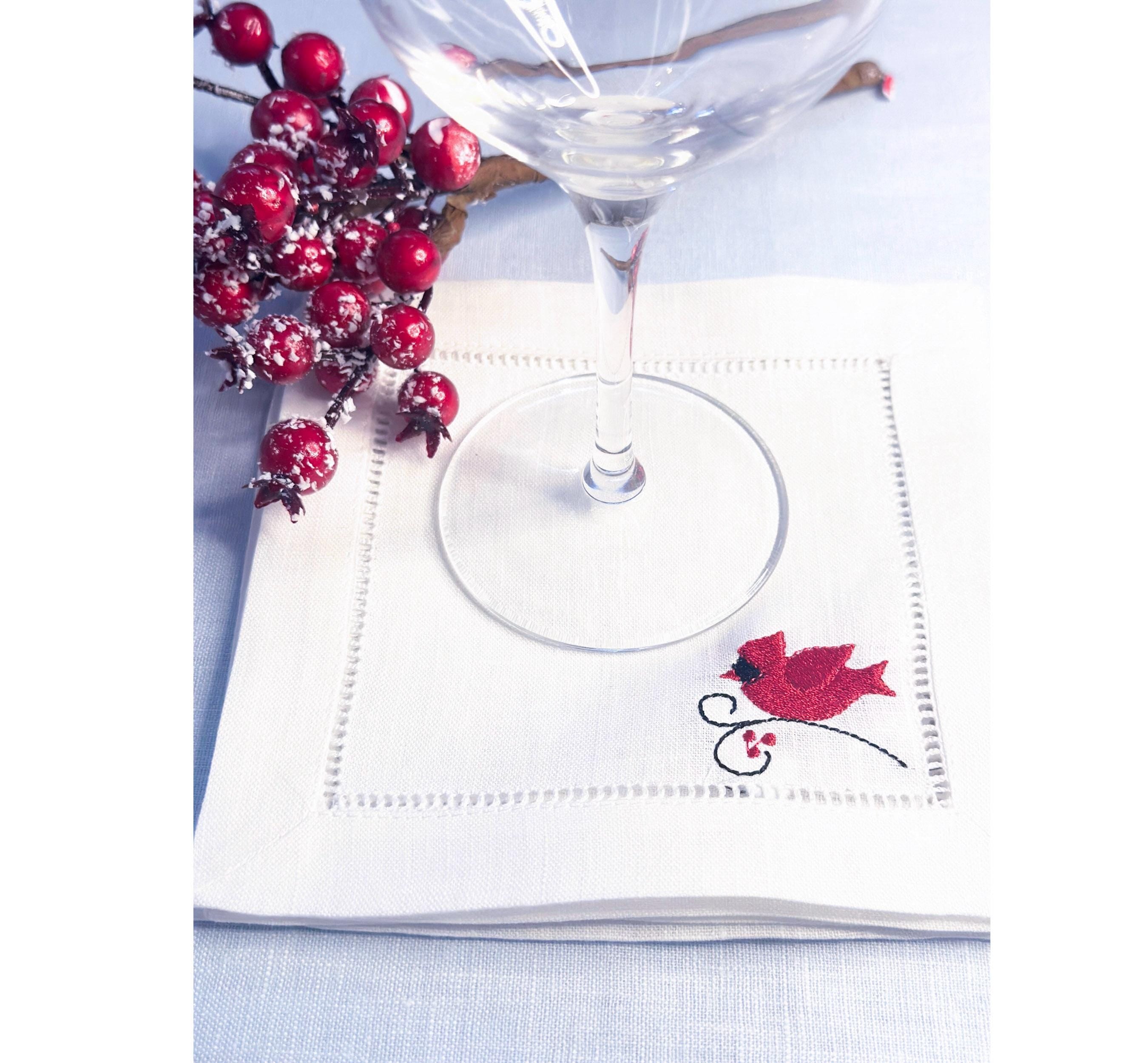 White linen cocktail napkin with a red male cardinal embroidered in the bottom right corner of the hemstitch napkin set on a light blue linen tablecloth with Christmas berries for bird lover gift.