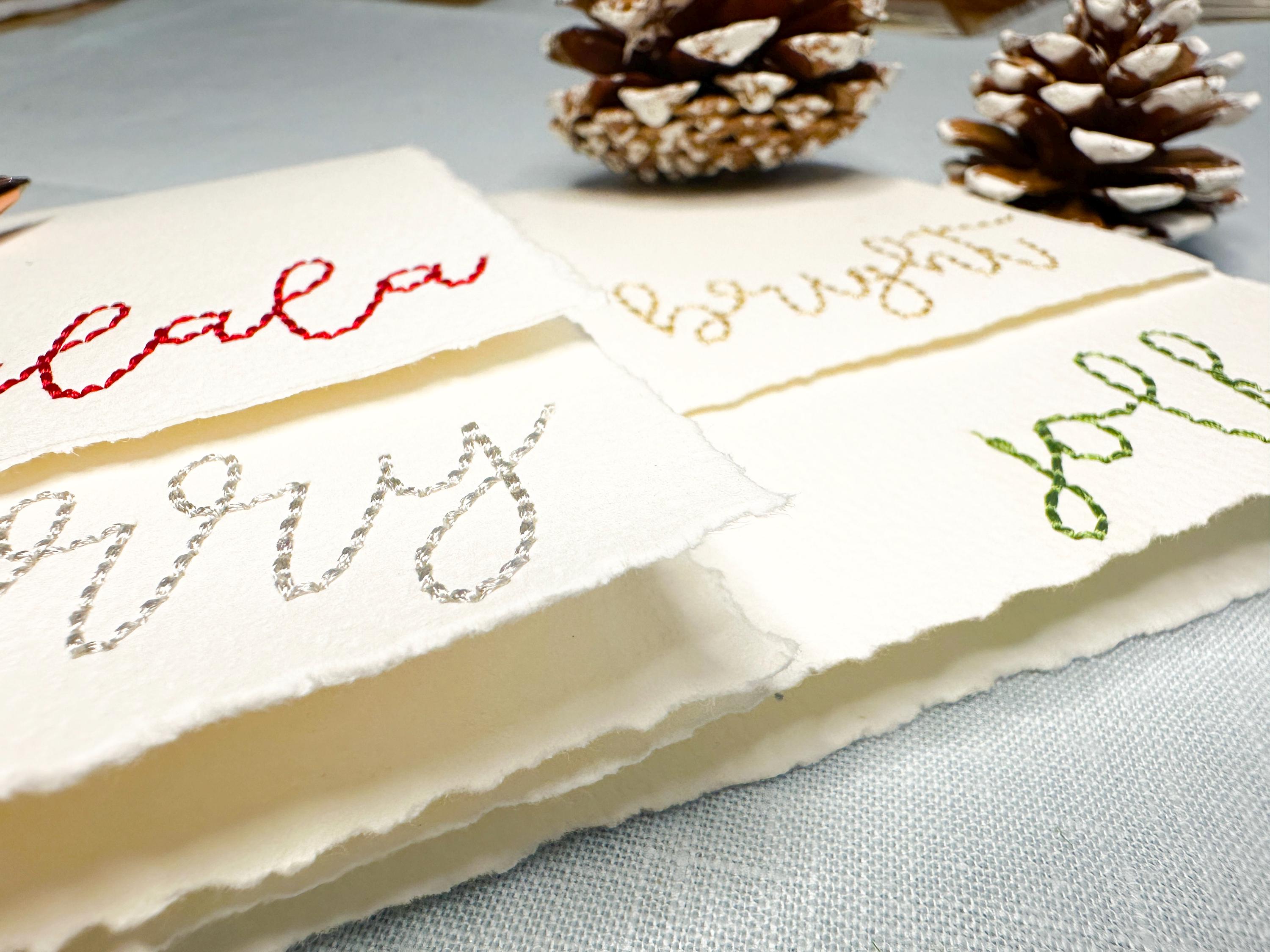Close up of holiday embroidered cards  stitched with a word in a different color: bright (in gold), merry (in silver), jolly (in green), and falala (in red) on a blue linen table cloths with pinecones and greenery. Perfect gift for school secretary.