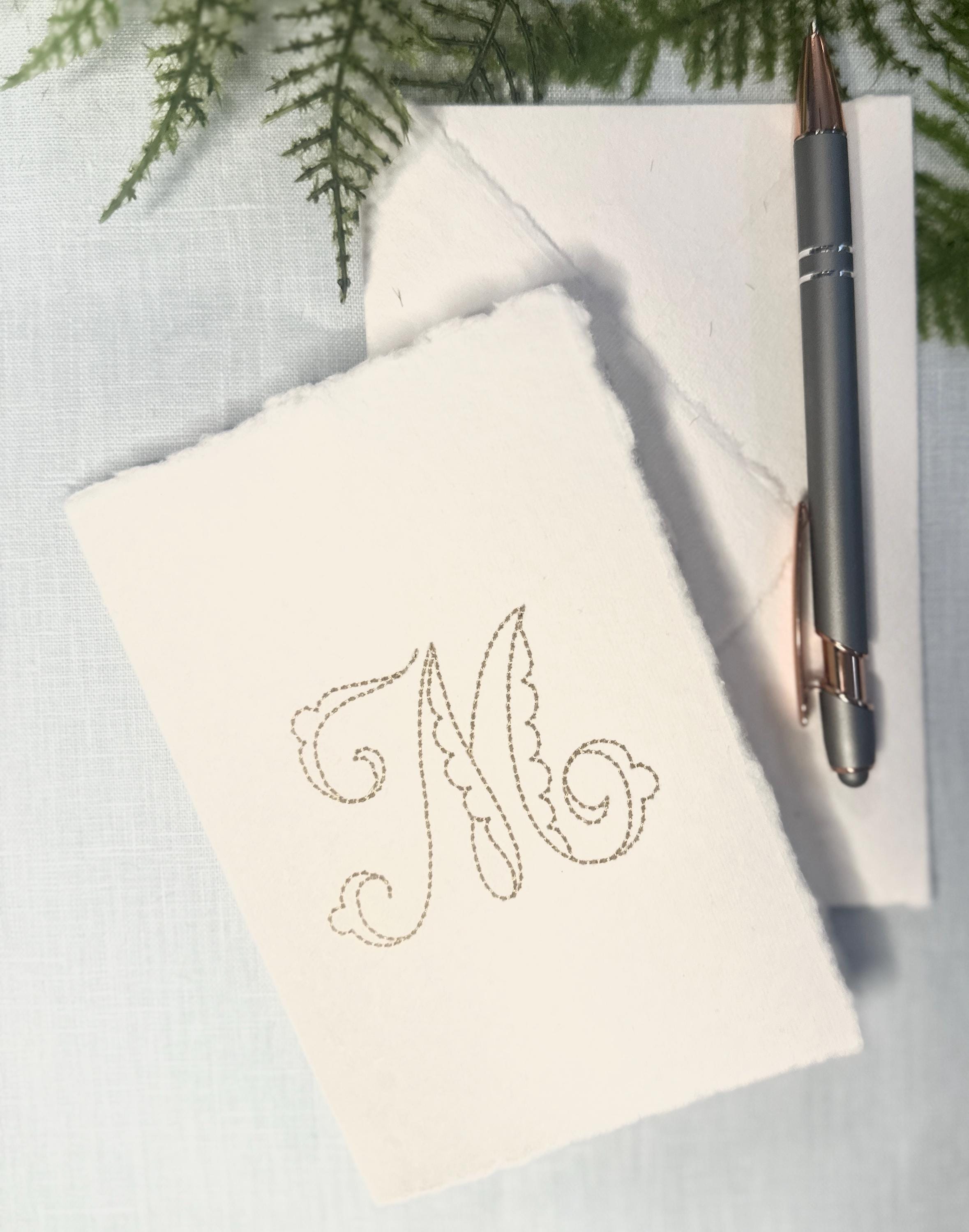 A card from a personalized stationary set of 4 note cards set on a blue linen background highlighting the delicate aesthetic edges of this custom stationary. The embroidery is in gold.
