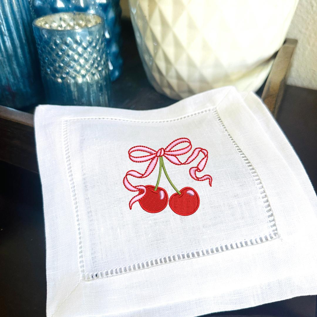 Set of 4 cherry embroidered linen cocktail napkins gift idea for 50th birthday gift for women. Two cherries on stem with a pink bow embroidered on a cocktail napkin and set on a bar with votive holders.