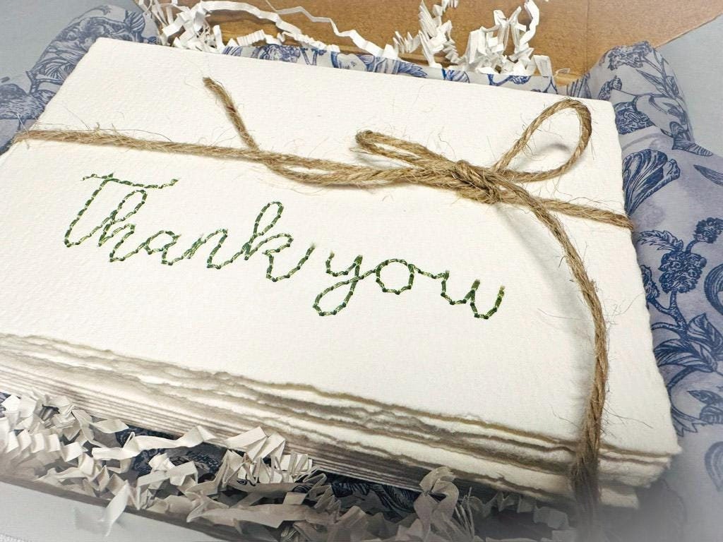 Pack of four thank you note cards packaged in a box and tied with jute string. The stitching is done in green and the cards are set in a box with white crinkle paper and blue and white decorated tissue.