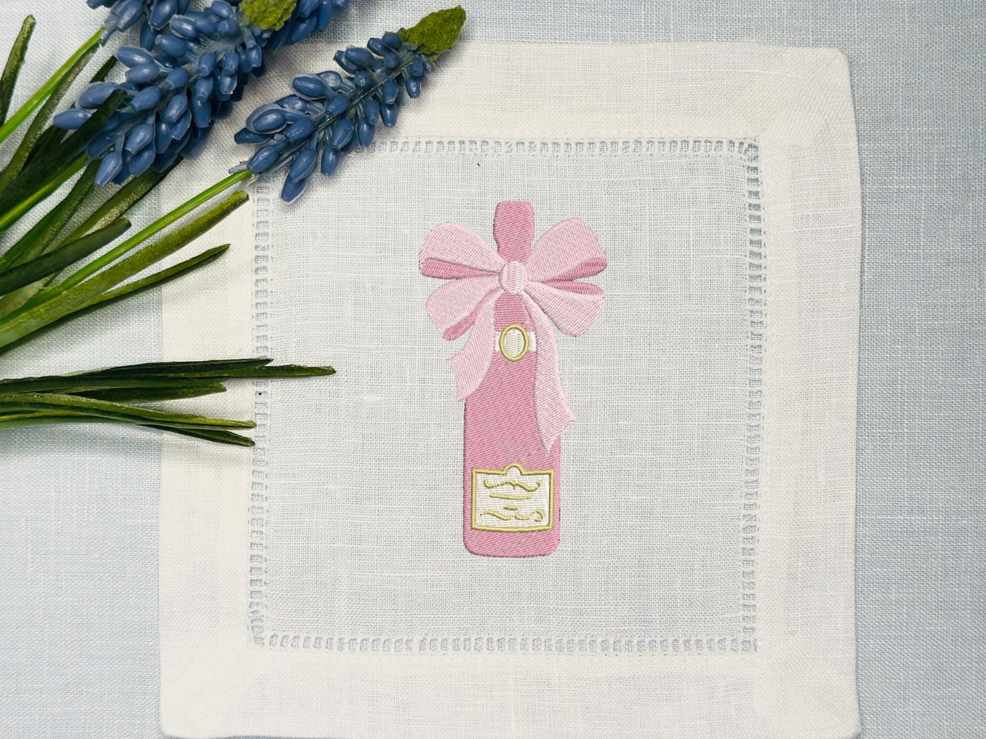Pink champagne bottle  with a bow embroidered on a simple white cocktail napkin for bachelorette party favor. Set upon a light blue linen tablecloth with blue flowers.