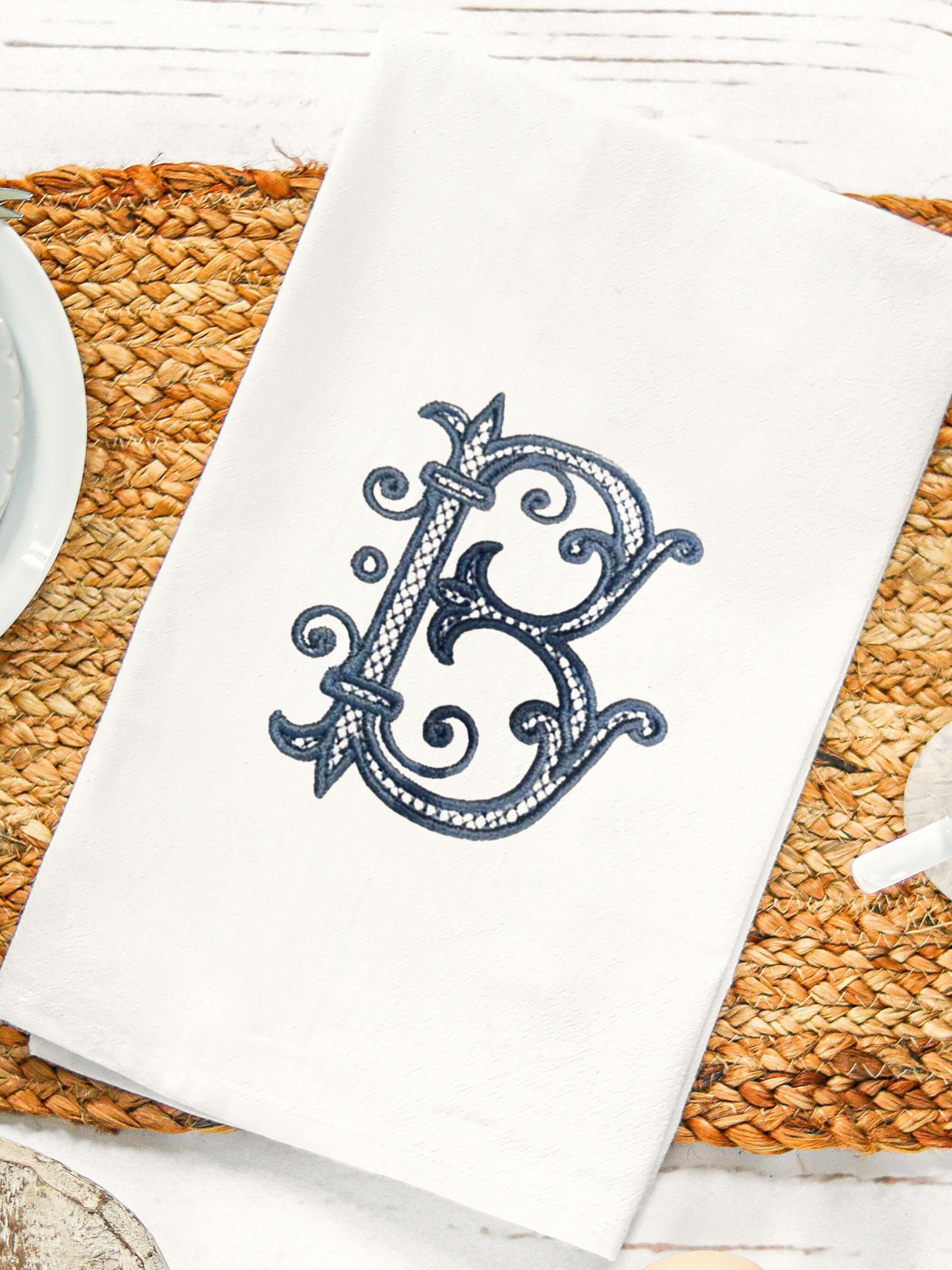 A white kitchen towel with a blue embroidered letter B in a decorative script font. Embroidered on a white tea towel set on a white board table with a jute tablecloth.