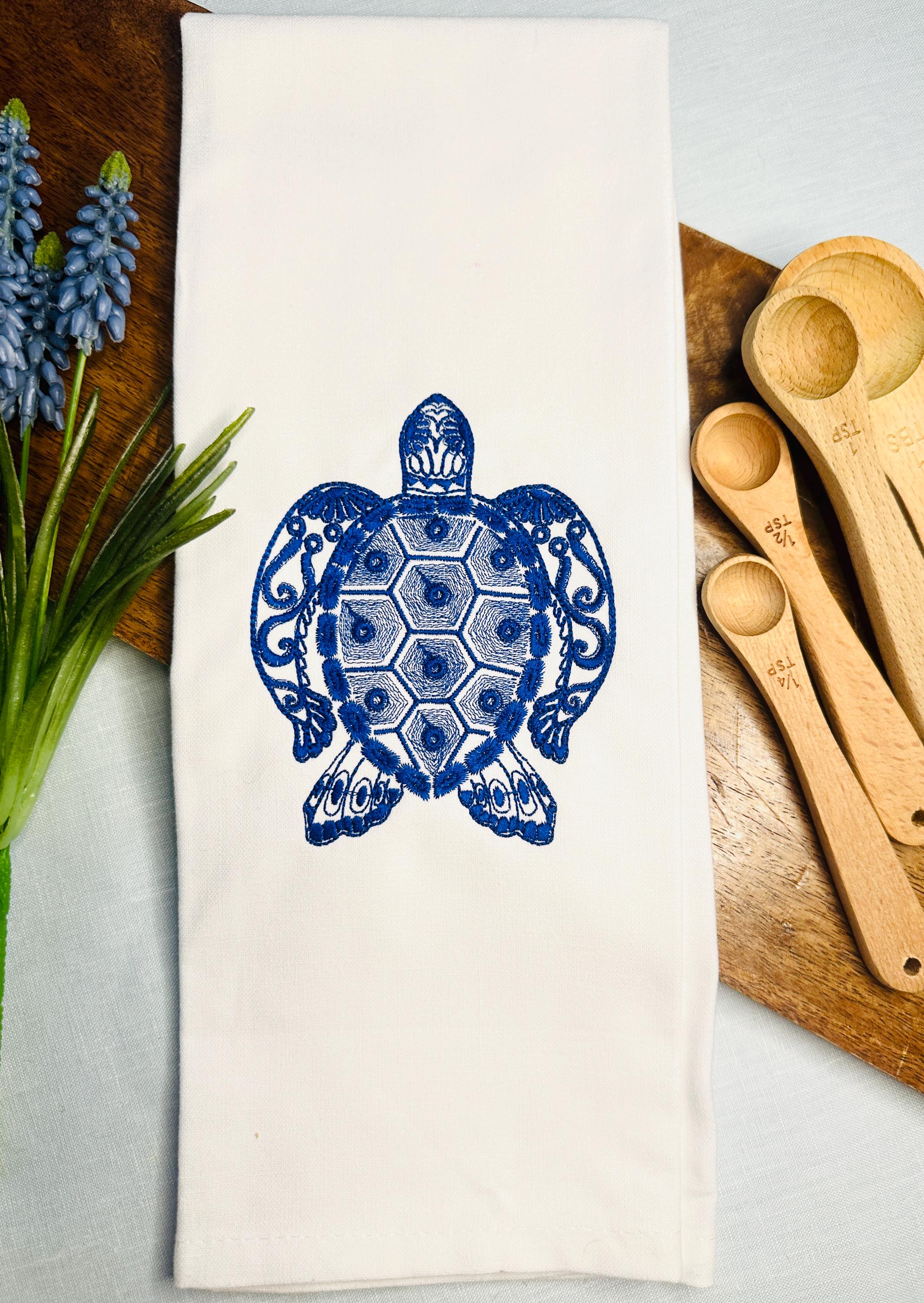 A blue sea turtle ocean themed gift embroidered on a white tea towel set on a wooden cutting board with flowers and measuring spoons.