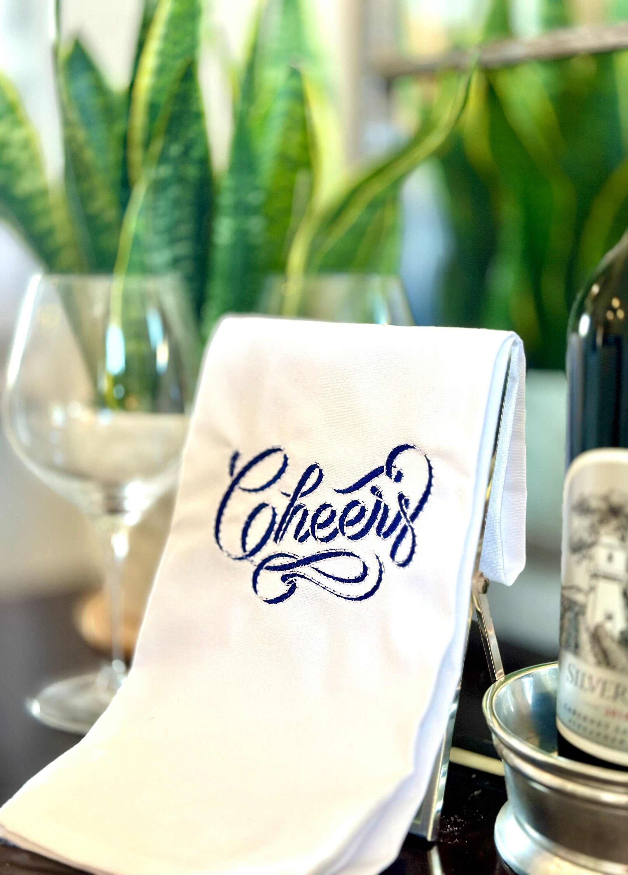 Cheers stitched in blue and white on a white tea towel for a 21st birthday gift for your sister! Set on a sidebar next to wine and wine glasses and a plant in the background.