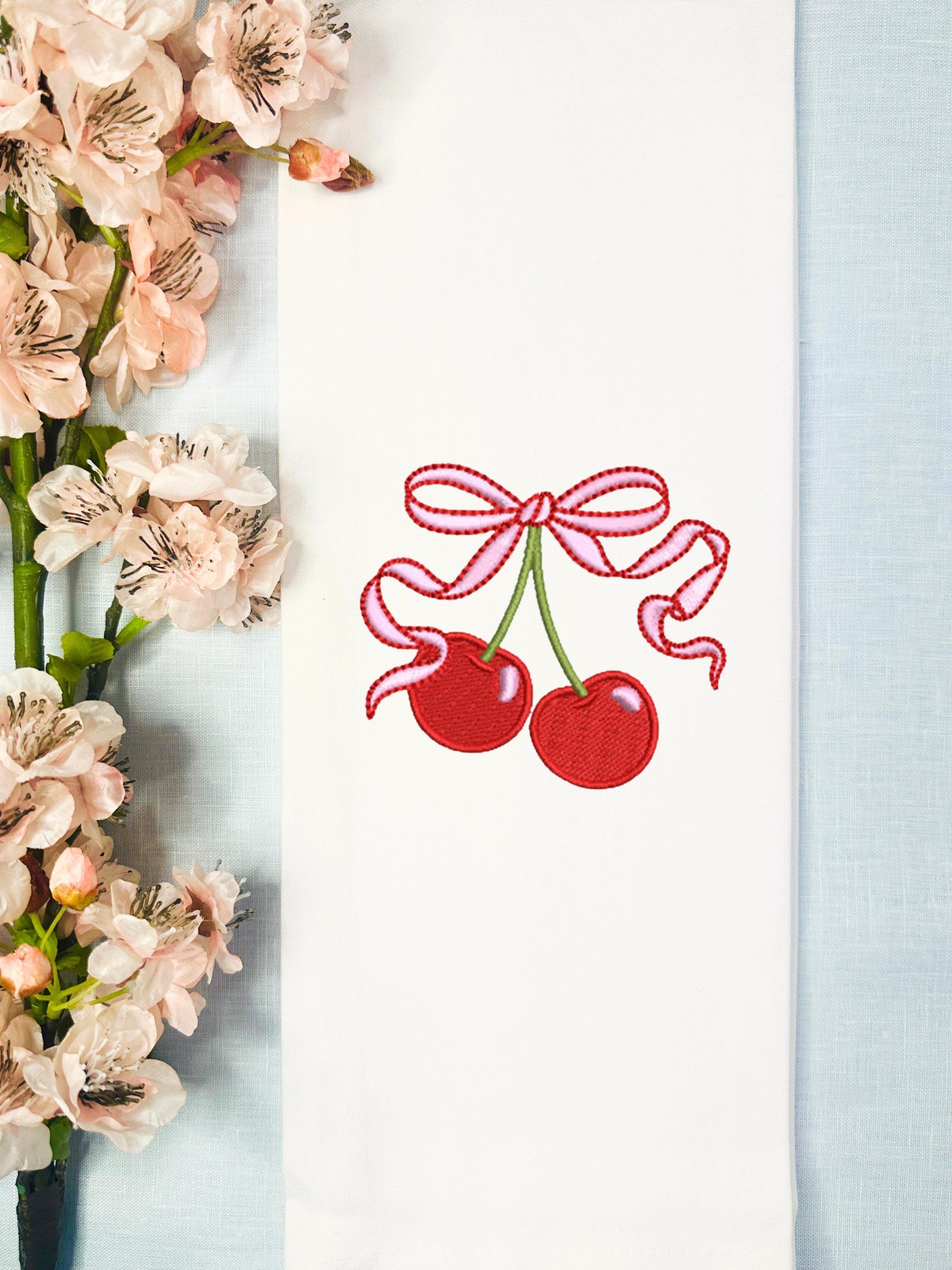 Embroidered red cherries with a pink bow set on a light blue linen tablecloth with cherry blossoms to the right.