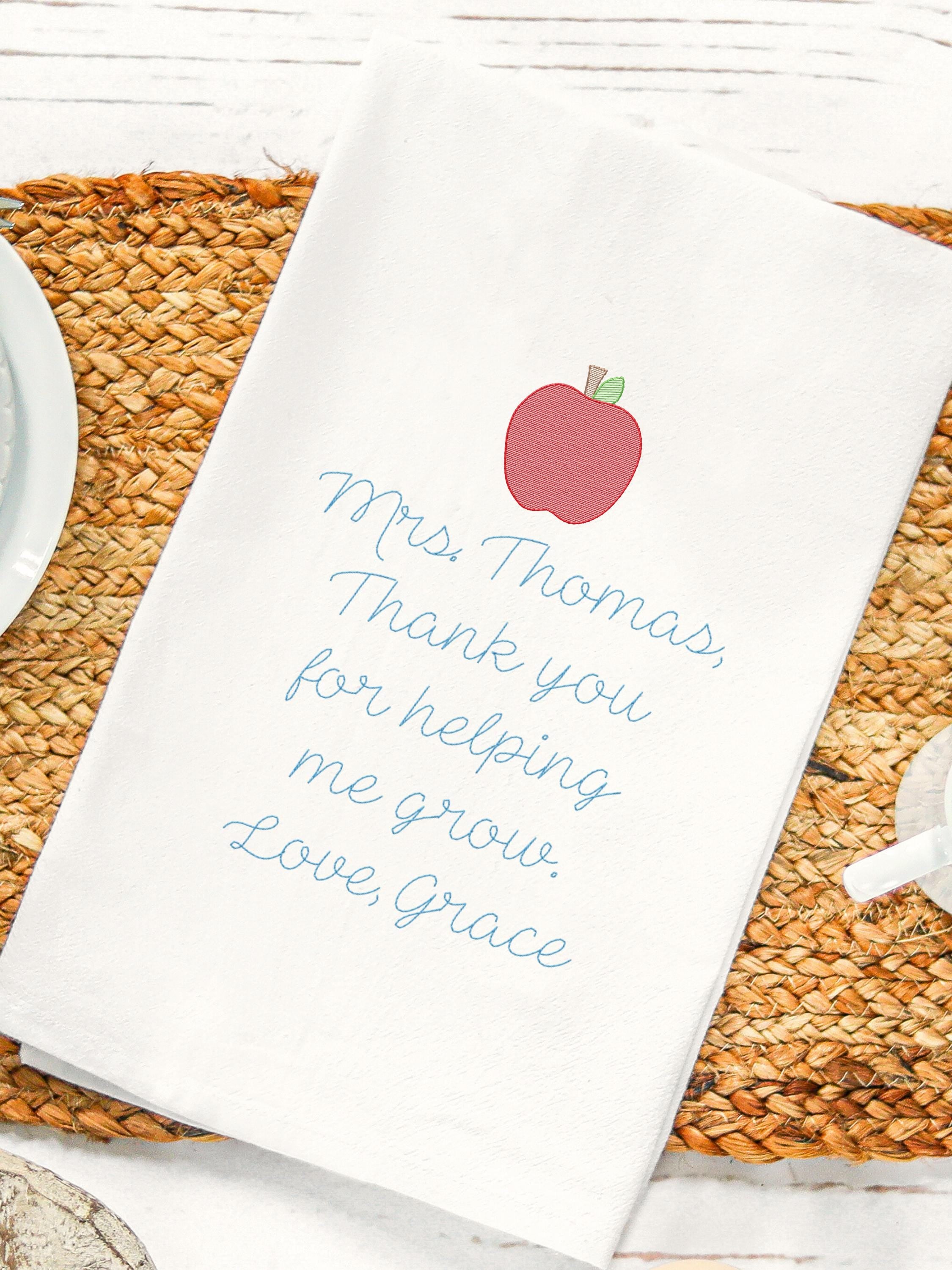 Teacher appreciation gift of an embroidered tea towel with a message that says: Mrs. Thomas, Thank you for helping me grow. Love, Grace. There is an apple on top of the message, also embroidered. It is set on a jute placemat with coffee cup & saucer.