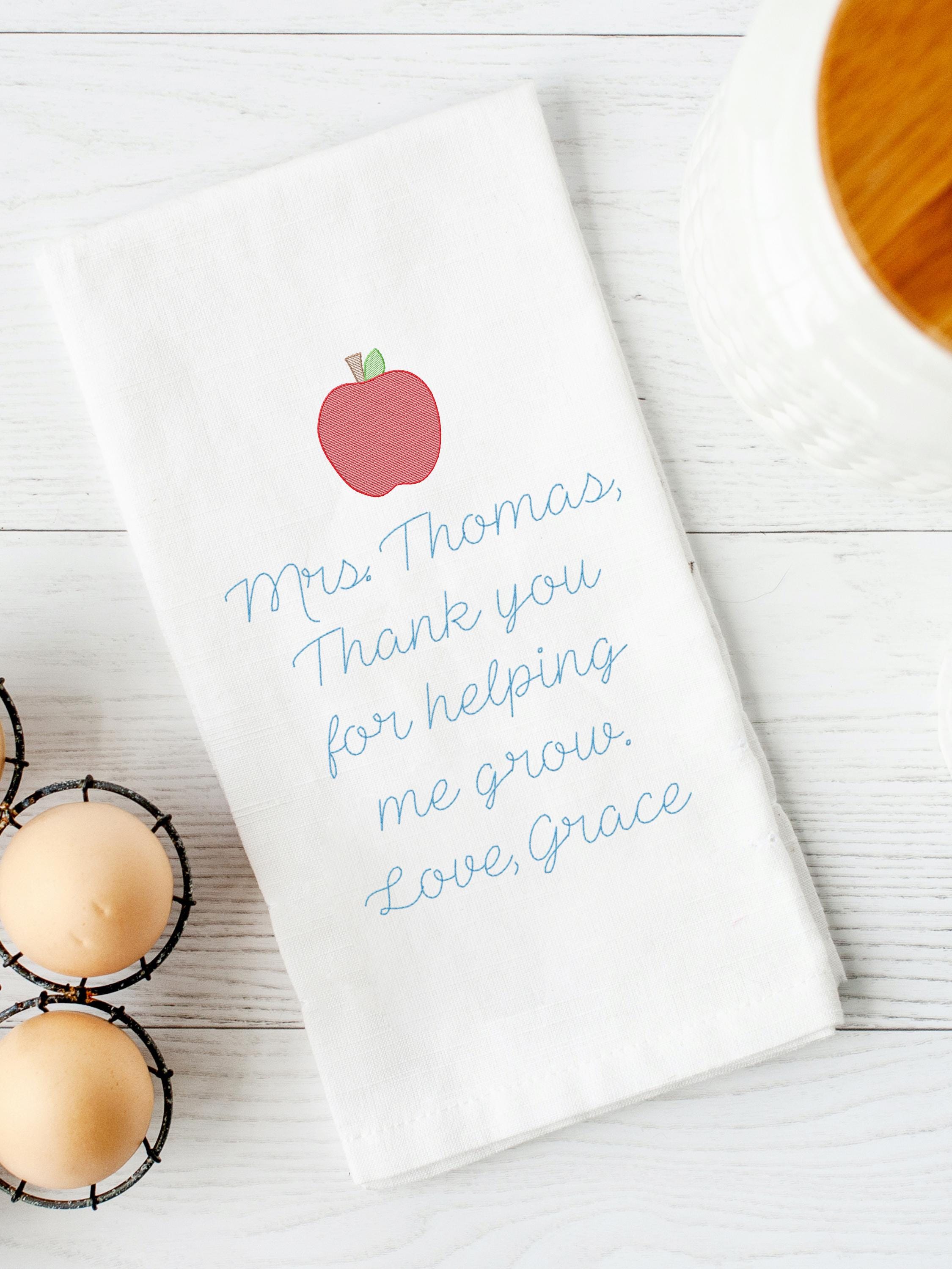 A white kitchen towel embroidered with a red apple and blue text that reads 'Mrs. Thomas, Thank you for helping me grow. Love, Grace.' The towel is set on a white board table next to eggs and a white canister with a wooden top.