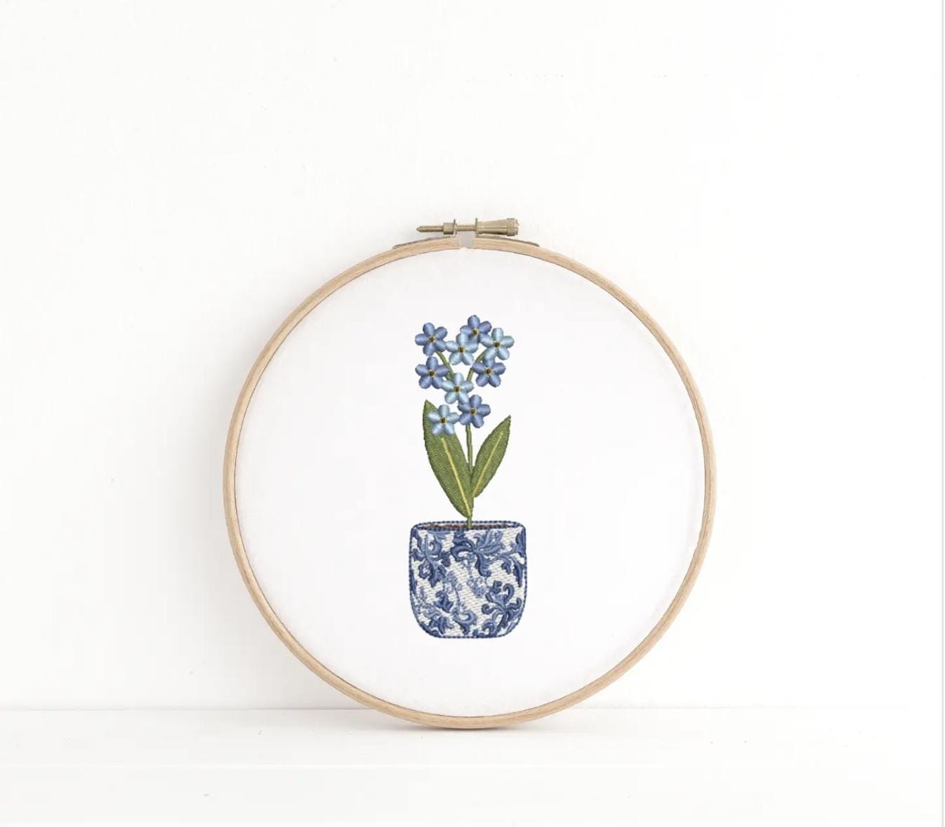 The chinoiserie coquette style pot of flowers embroidered on a white background in a wooden embroidery hoop.