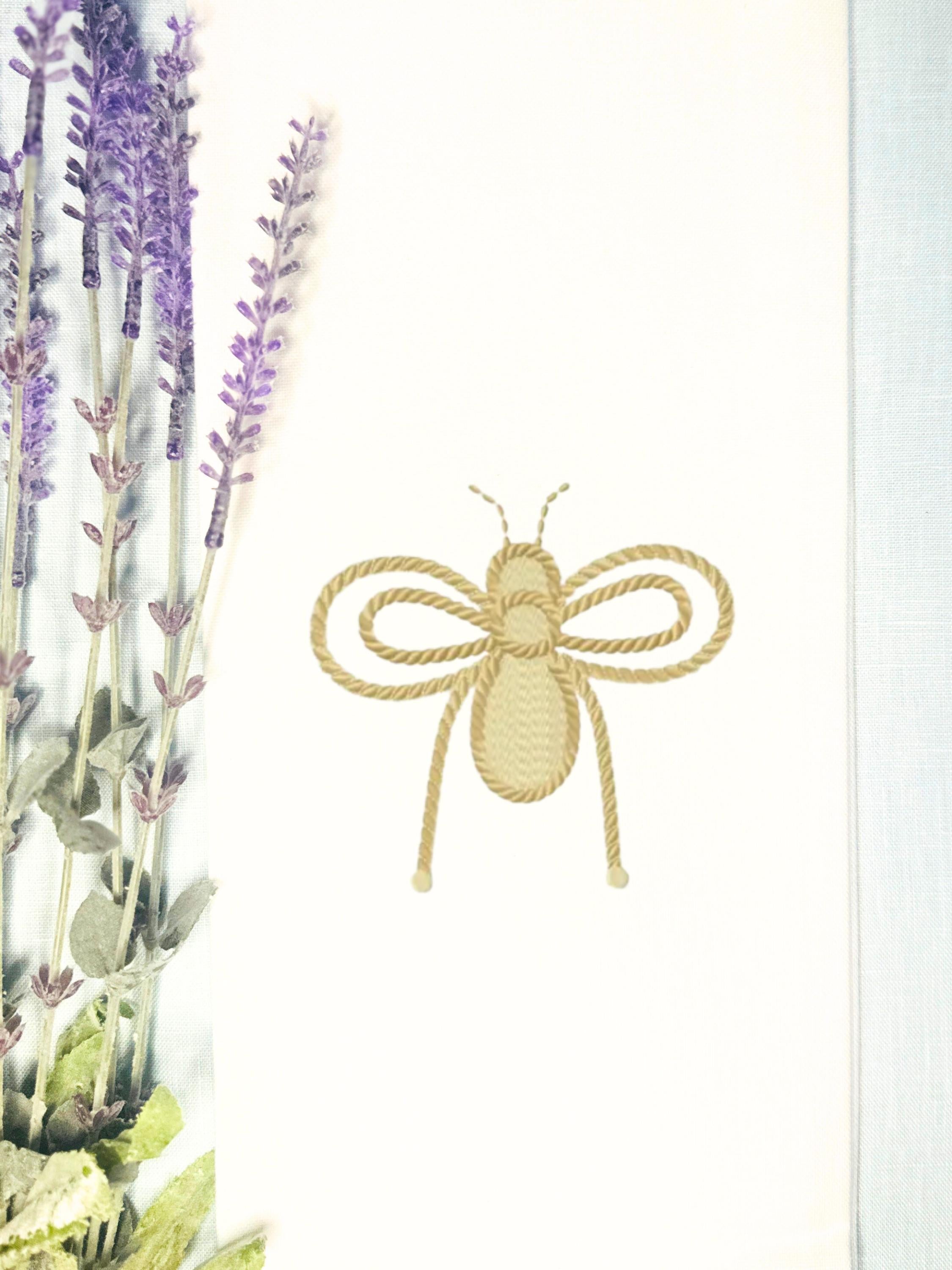 A honey bee embroidered in gold on a white tea towel on a light blue linen tablecloth with lavender next to it.