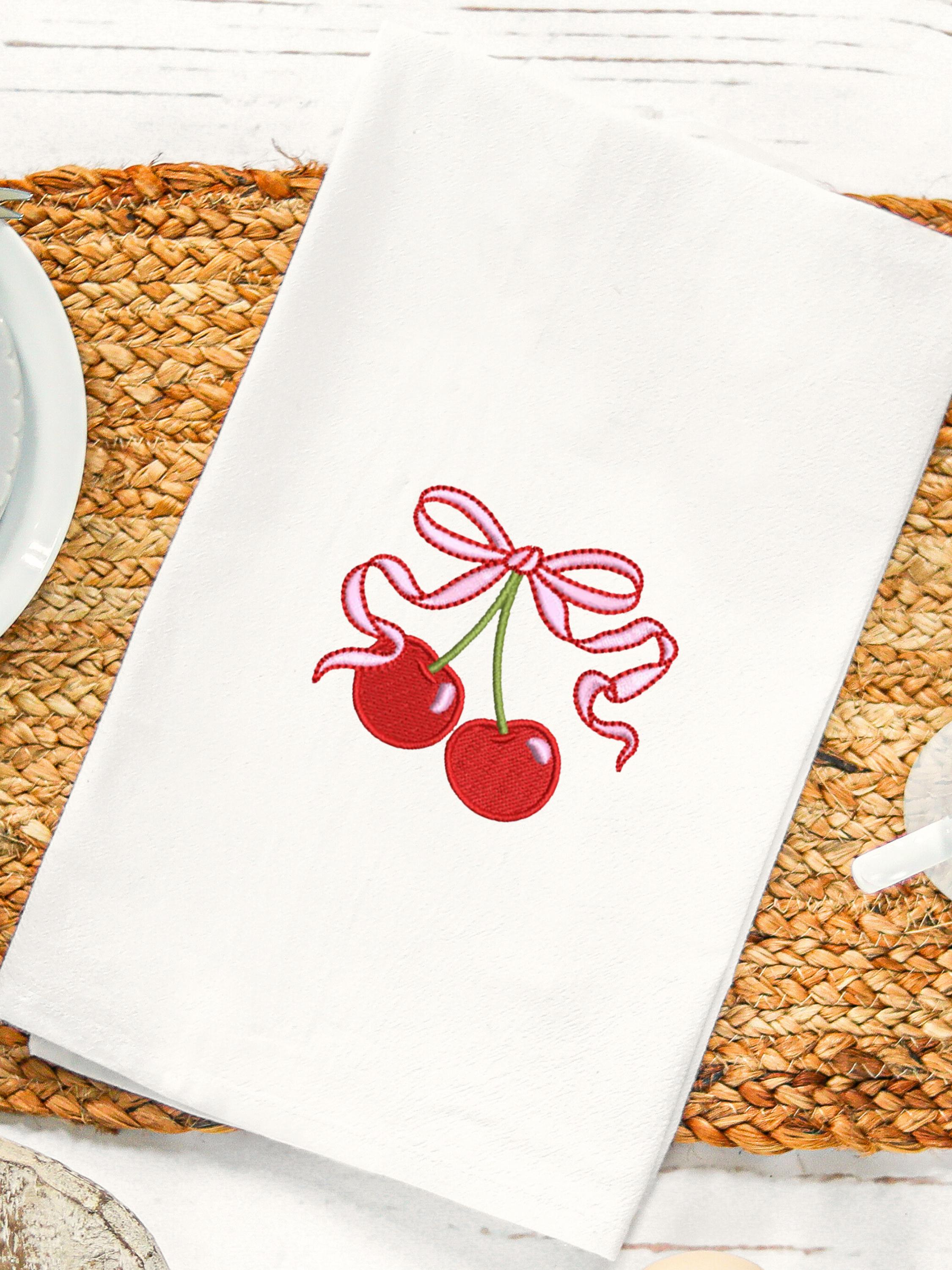 A white tea towel with red cherries tied in a bow laid on a wicker placemat on a white table with white dishware.