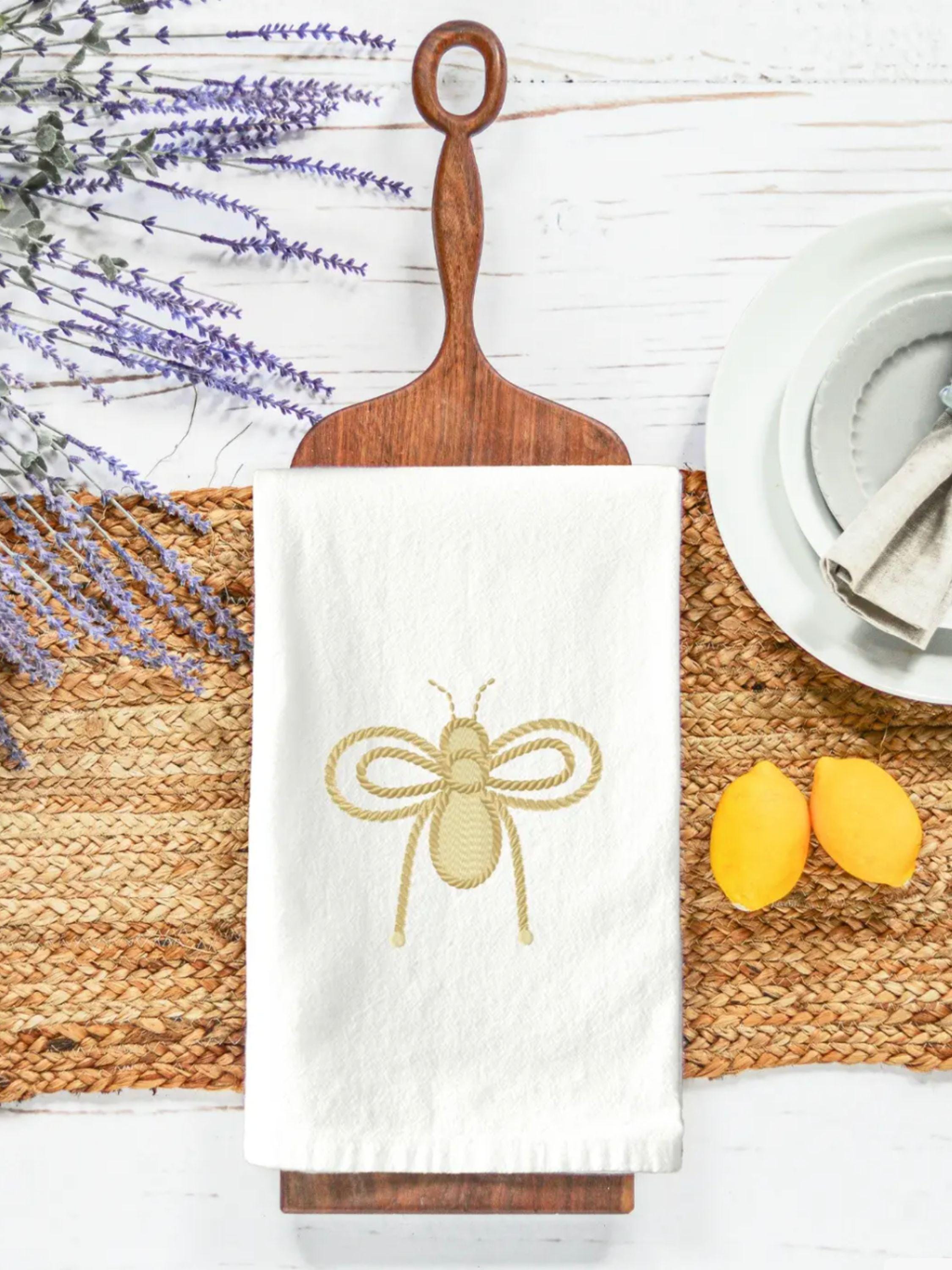 Bee kitchen decor of a honey bee with rope design embroidered on a white kitchen towel with lemons and plates to the right, the towel set on a decorative cutting board on a table.