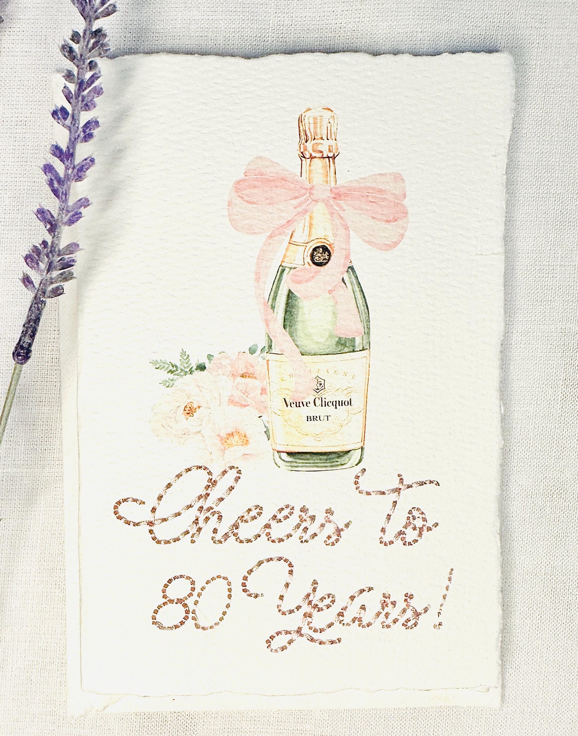 A birthday card with a watercolor champagne bottle with a pink bow and flowers to the left side. Under the art is an embroidered message: Cheers to 80 Years in pink thread. The card and matching envelope are set on a white linen tablecloth with a sprig of lavender to the left of the card.