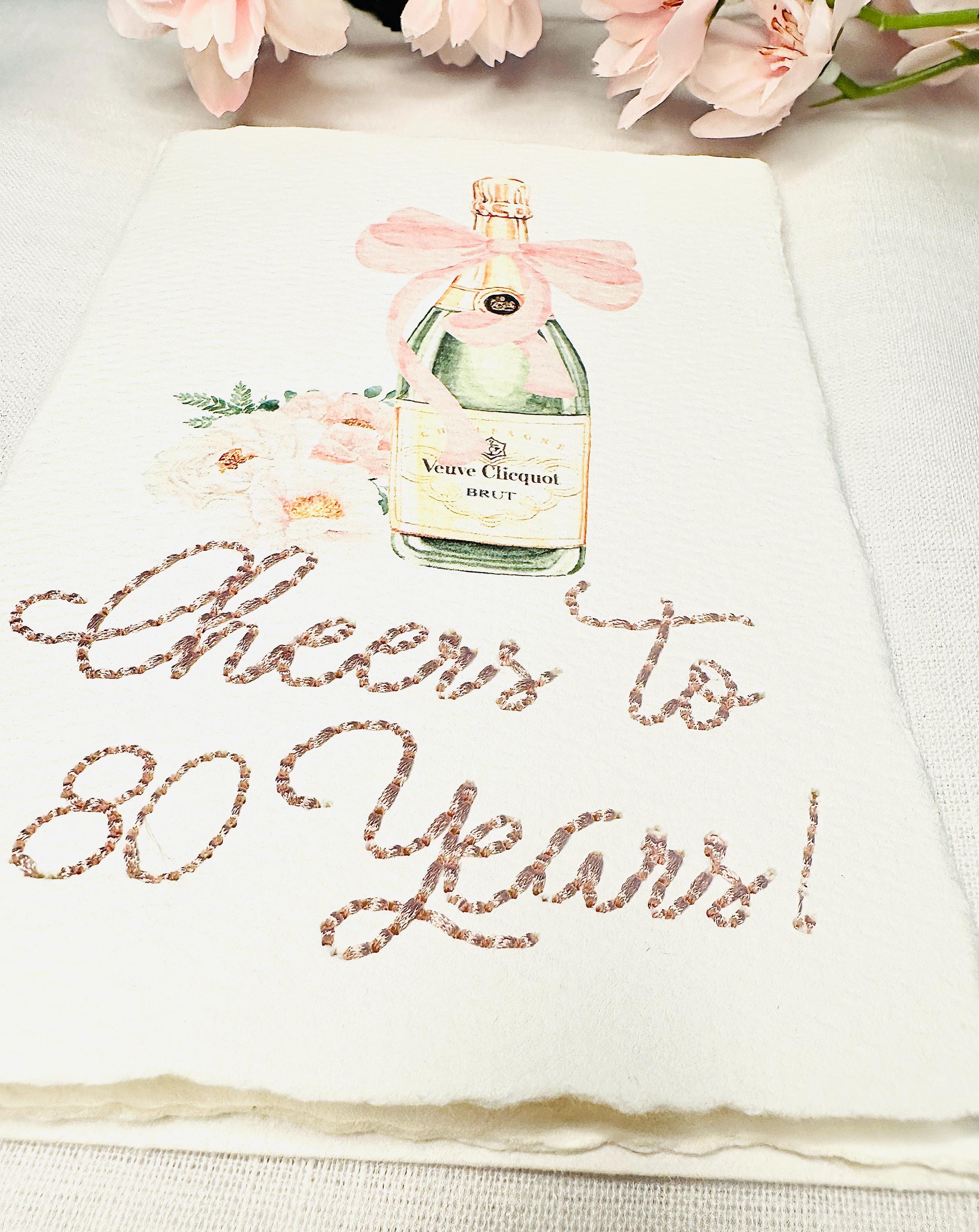 A birthday card with a watercolor champagne bottle with a pink bow and flowers to the left side. Under the art is an embroidered message: Cheers to 80 Years in pink thread. The card and matching envelope are set on a white linen tablecloth with pink cherry blossoms on top.